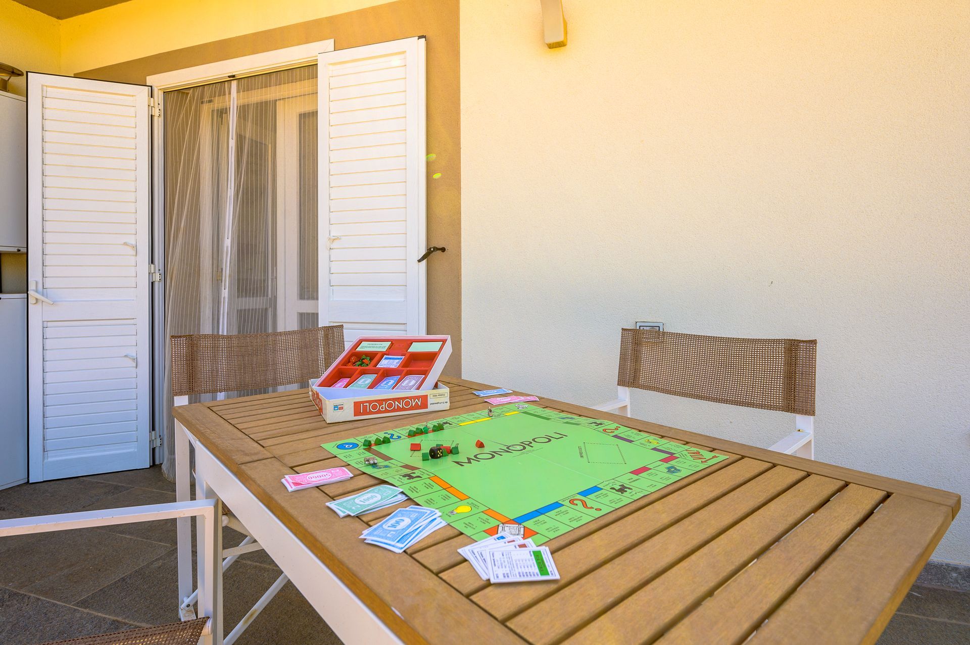 Gioco del Monopoli su un tavolo di legno all'aperto, vicino a persiane bianche aperte.