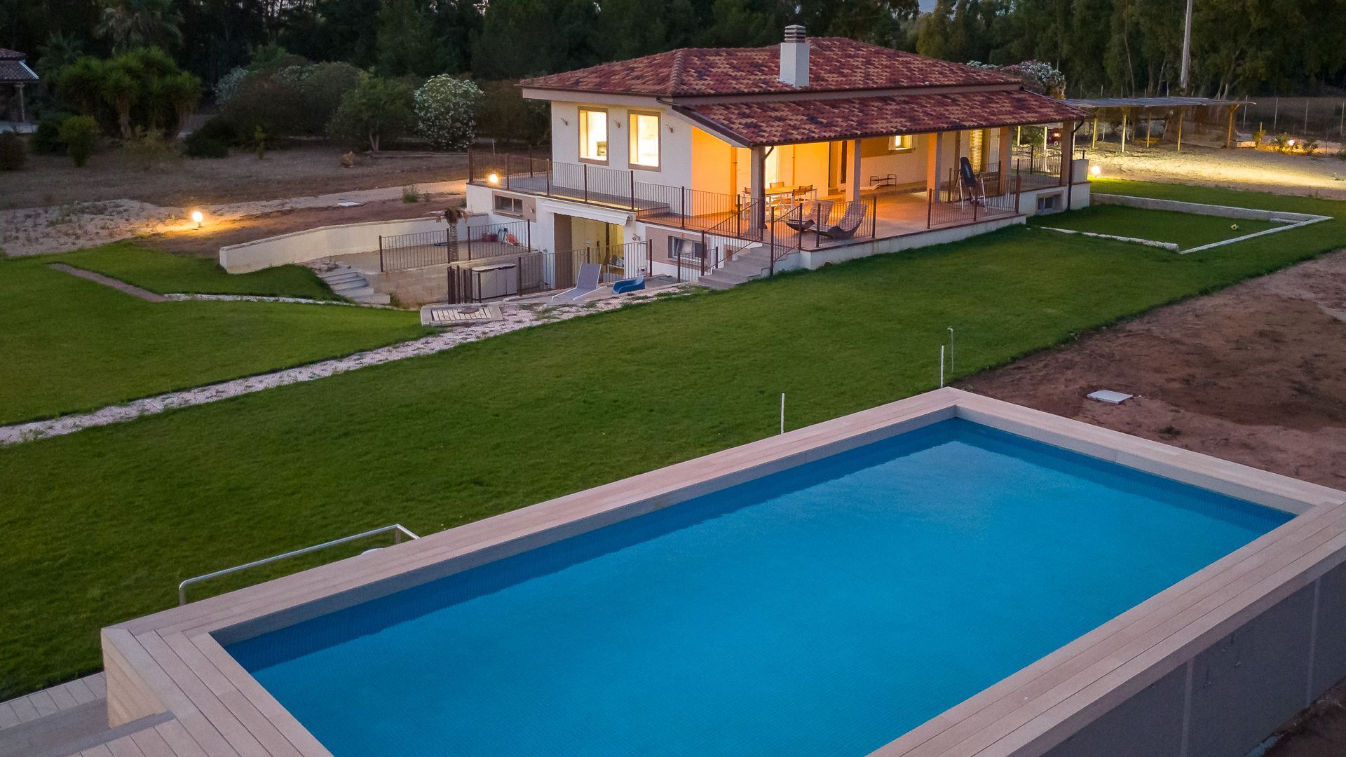 Casa con piscina. Casa a due piani, color crema, illuminata dall'interno, accanto a una piscina rettangolare blu su un prato erboso.