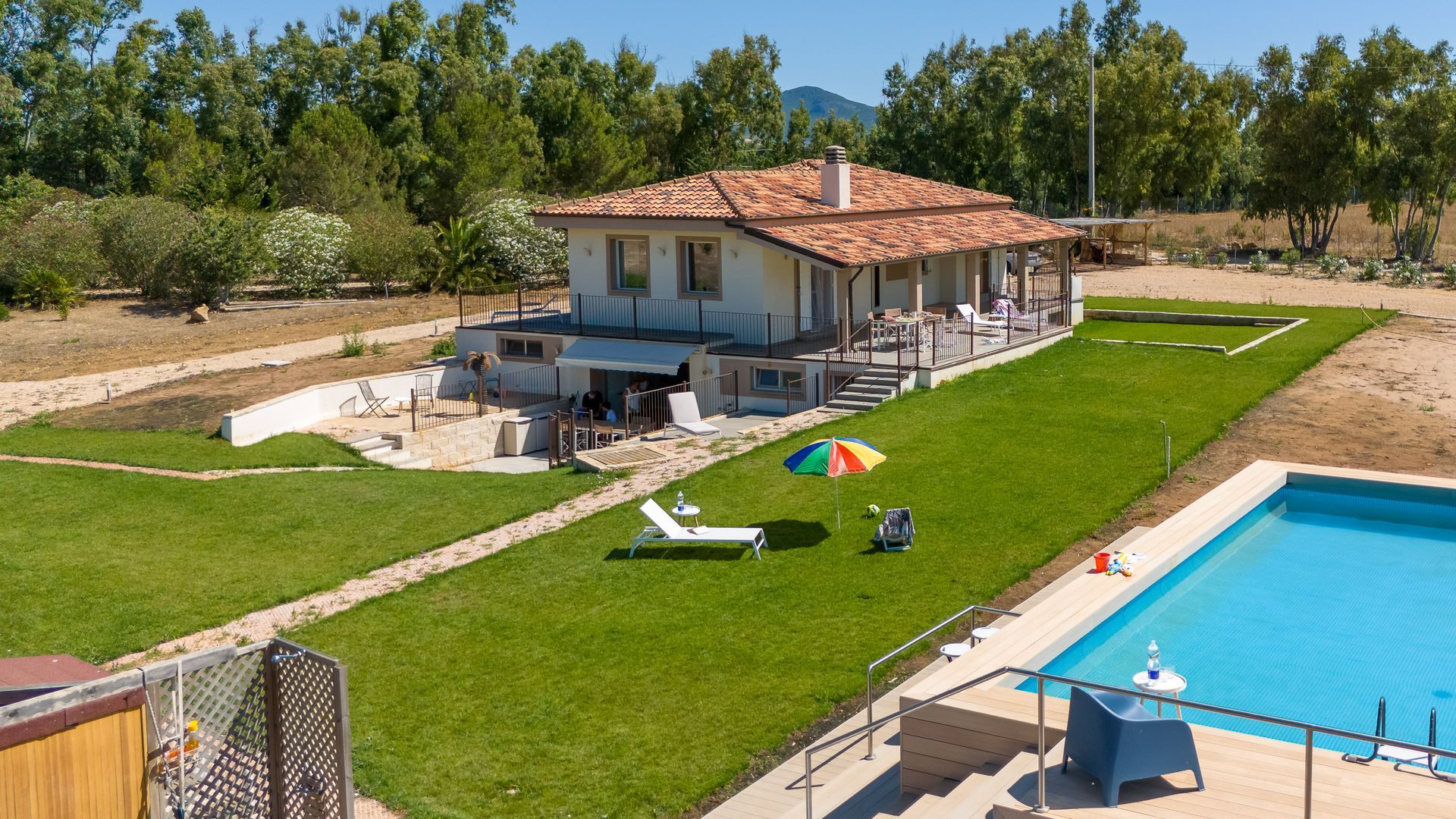Casa con piscina e prato verde in una giornata di sole. Una persona si rilassa su una sdraio sotto un ombrellone.