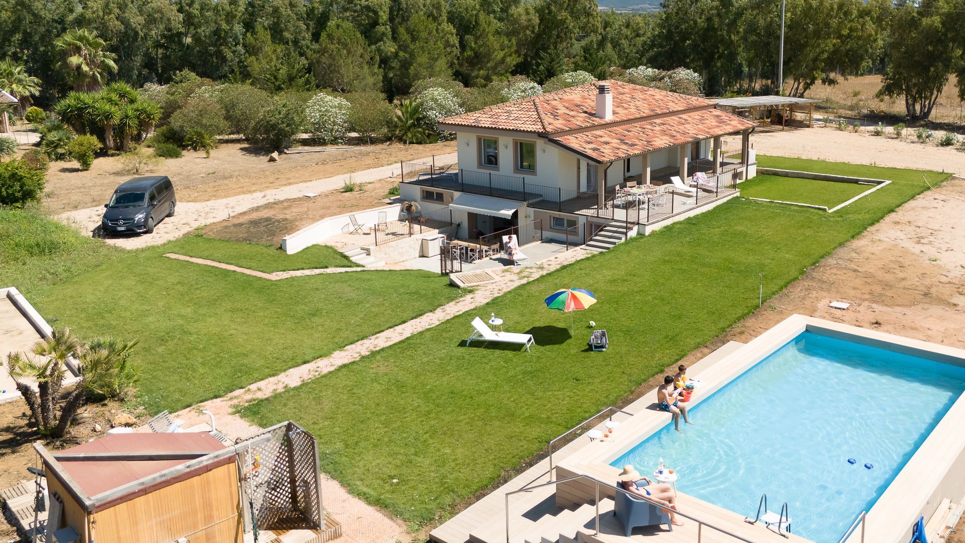 Casa con piscina e prato; gente che si rilassa. Furgone nero parcheggiato. Giornata di sole.