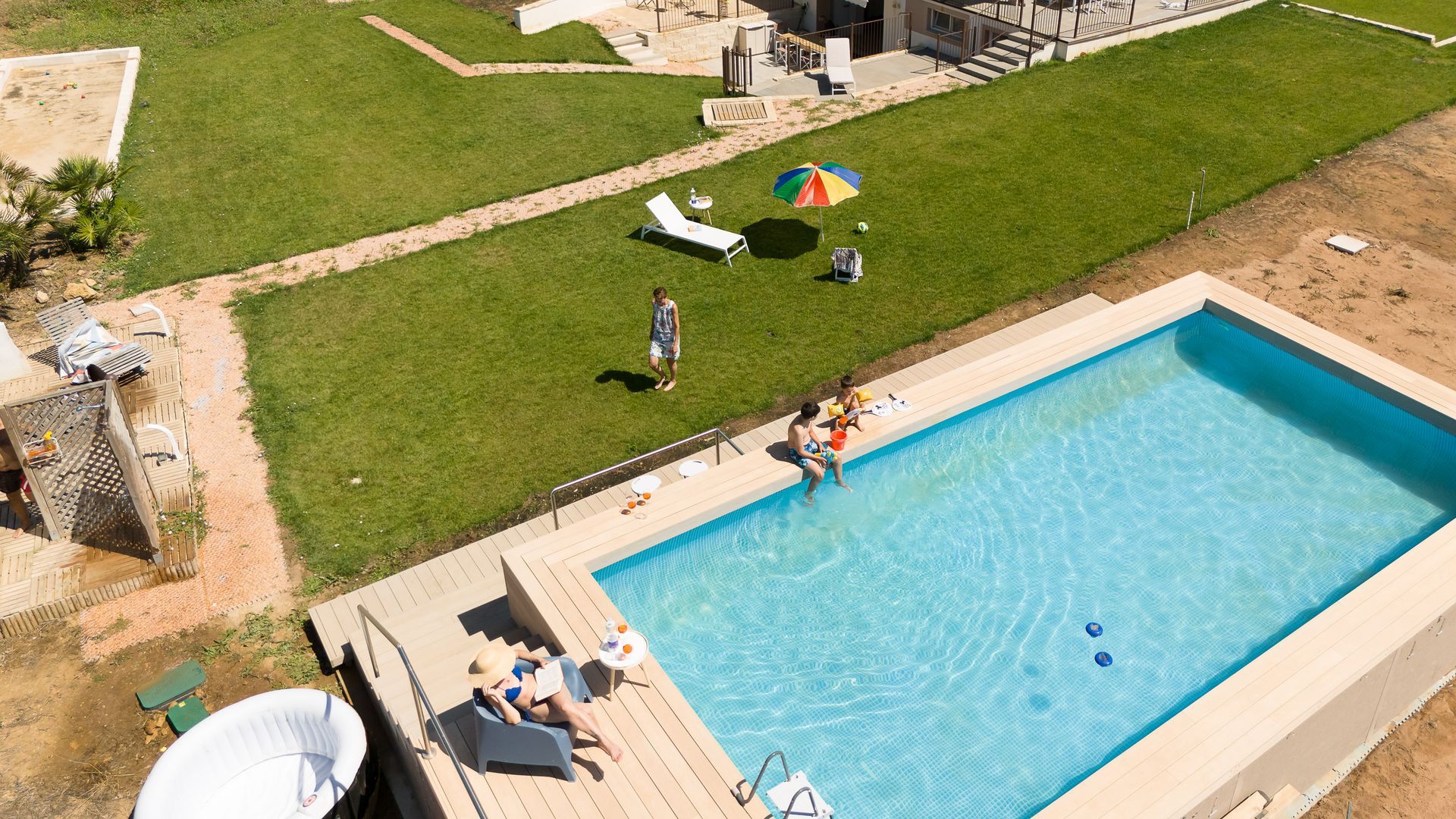 Persone che si rilassano a bordo piscina, tra cui bagnanti e persone sedute sul bordo, con un prato e un edificio nelle vicinanze.