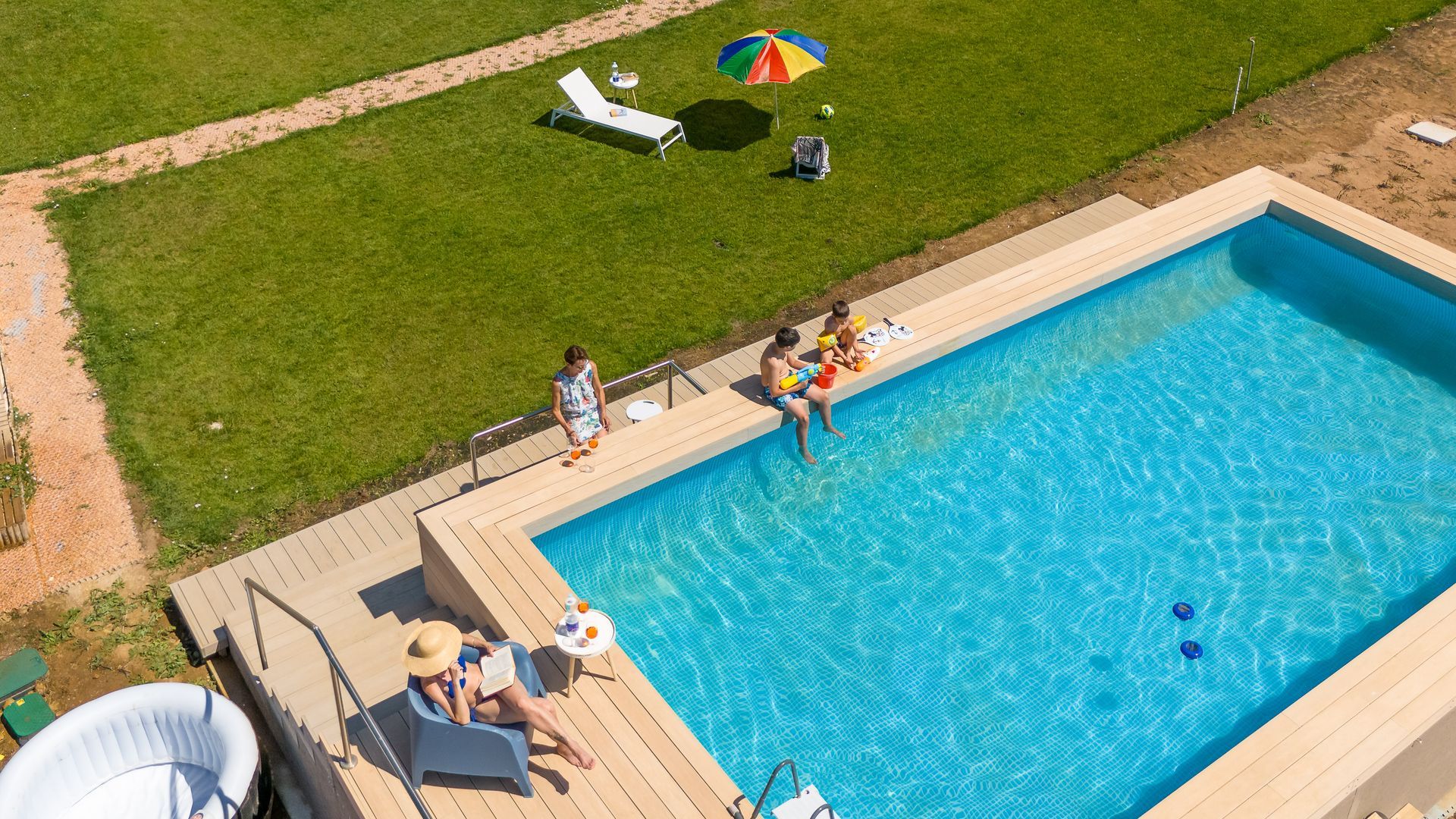 Persone che si rilassano a bordo di una piscina rettangolare con acqua azzurra, prato, ombrellone e jacuzzi.