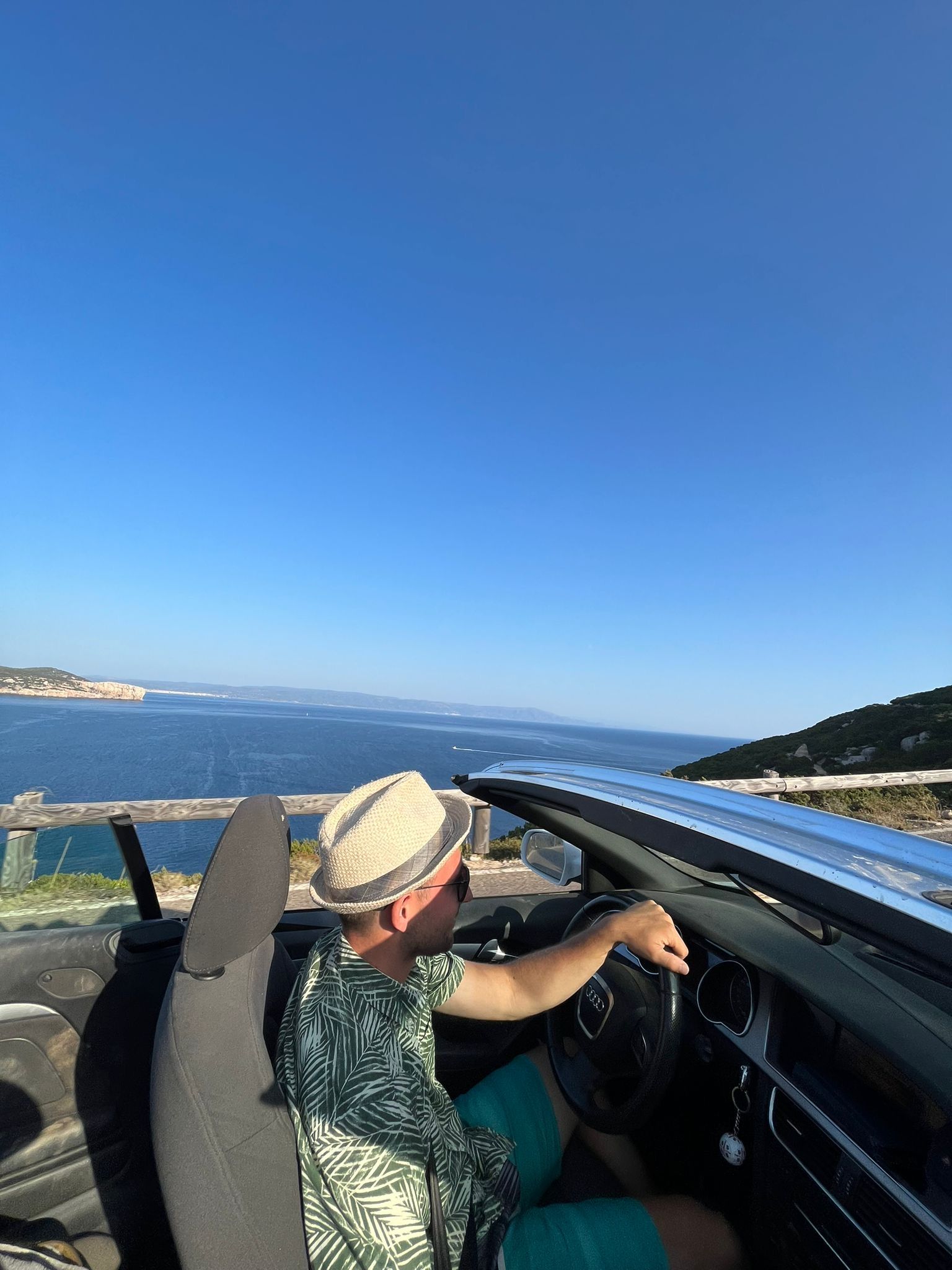 Un uomo in una decappottabile guida lungo una strada costiera. Acqua e cielo blu. Indossa un cappello e una camicia stampata.