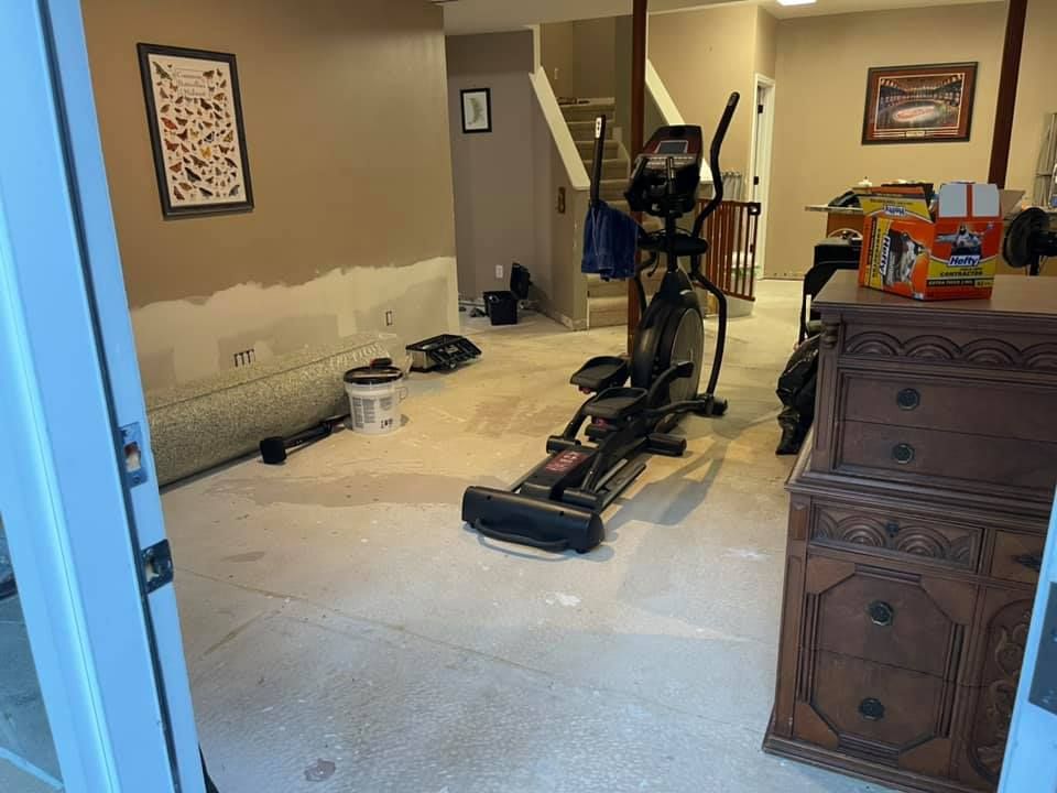 Basement undergoing renovation. Elliptical machine, furniture, and tools sit on the floor. Walls have exposed drywall.