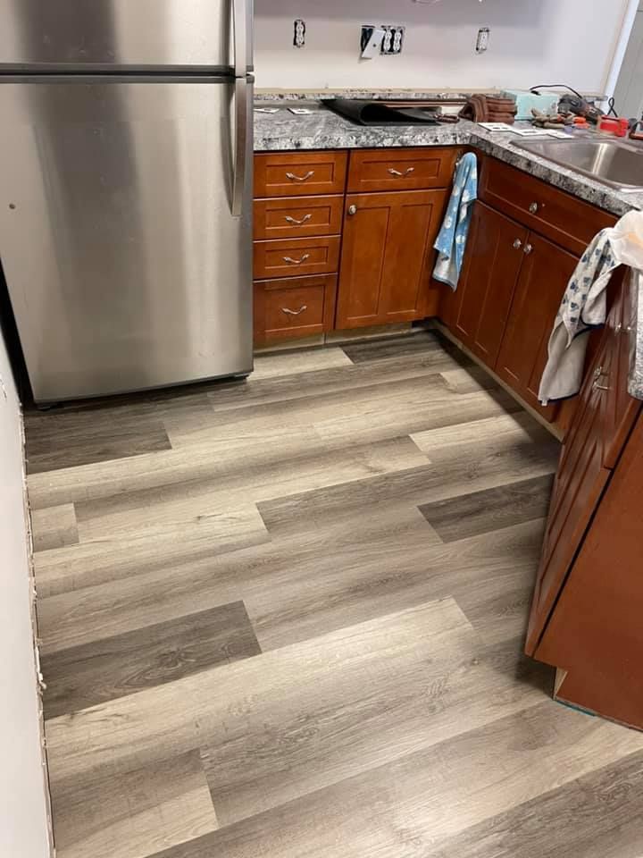 Kitchen with new grey wood-look flooring. Brown cabinets, stainless steel fridge, and countertop.