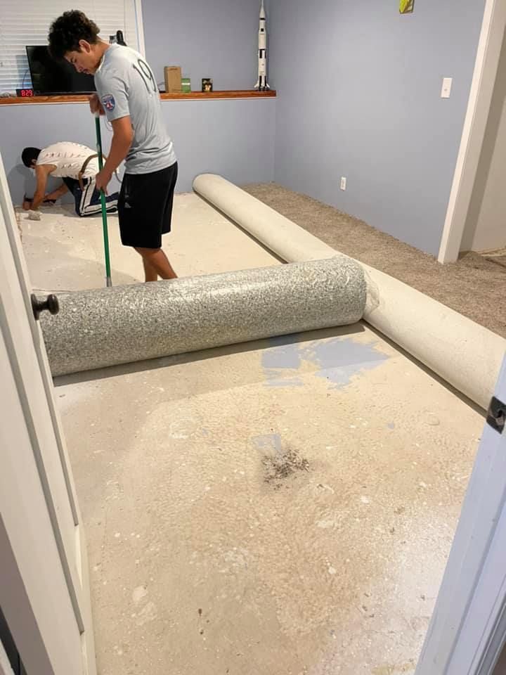 Two people rolling out carpet in a room with light blue walls and a concrete floor.