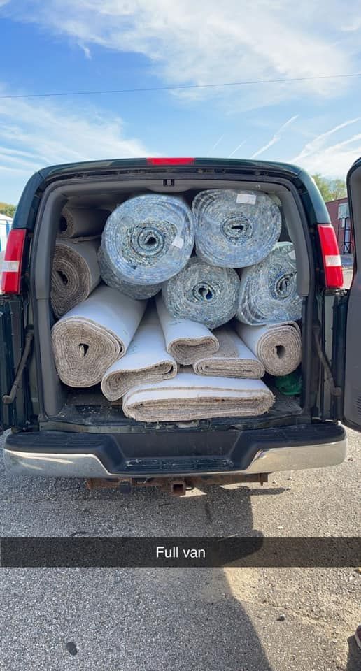 A van's cargo area is filled with rolled-up carpets of various sizes and colors, parked outdoors.