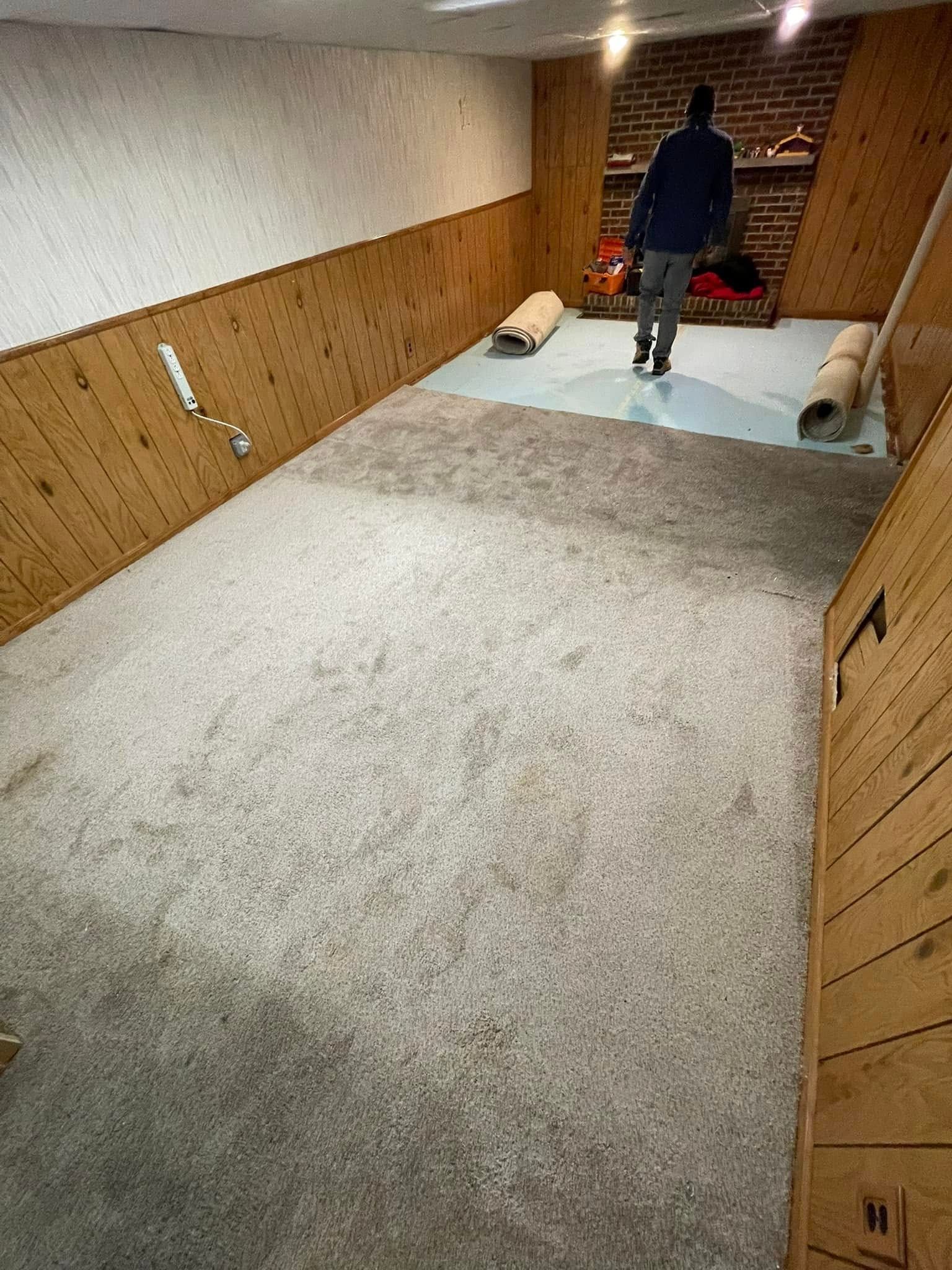 Person standing on a light gray carpeted floor in a room with wood paneling and a rolled-up rug.