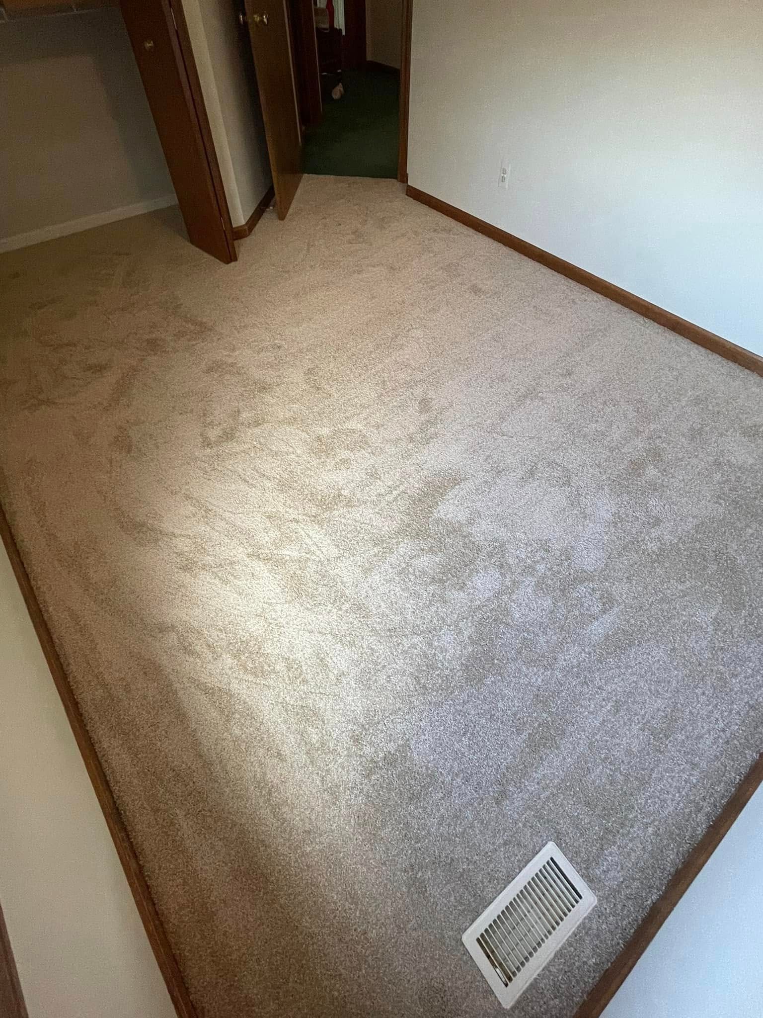 Beige carpeted room with brown trim and a vent. A door and part of a hallway are visible.