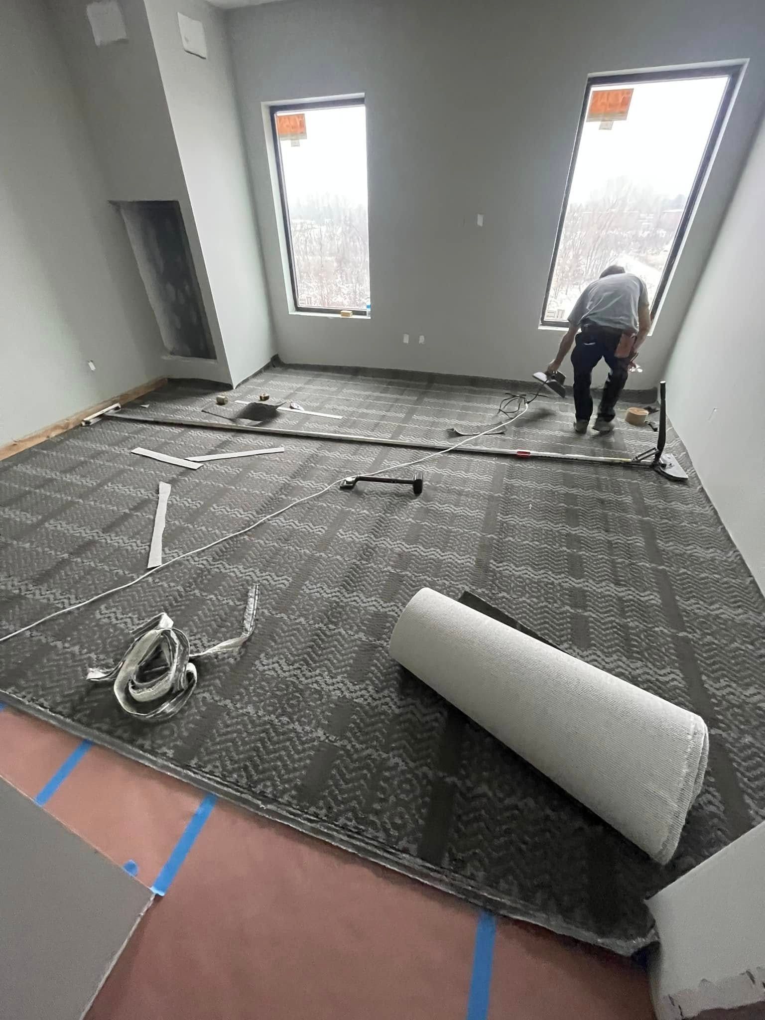 A person installing carpet tiles in a room with two windows. Gray and white carpet, with tools laid out.