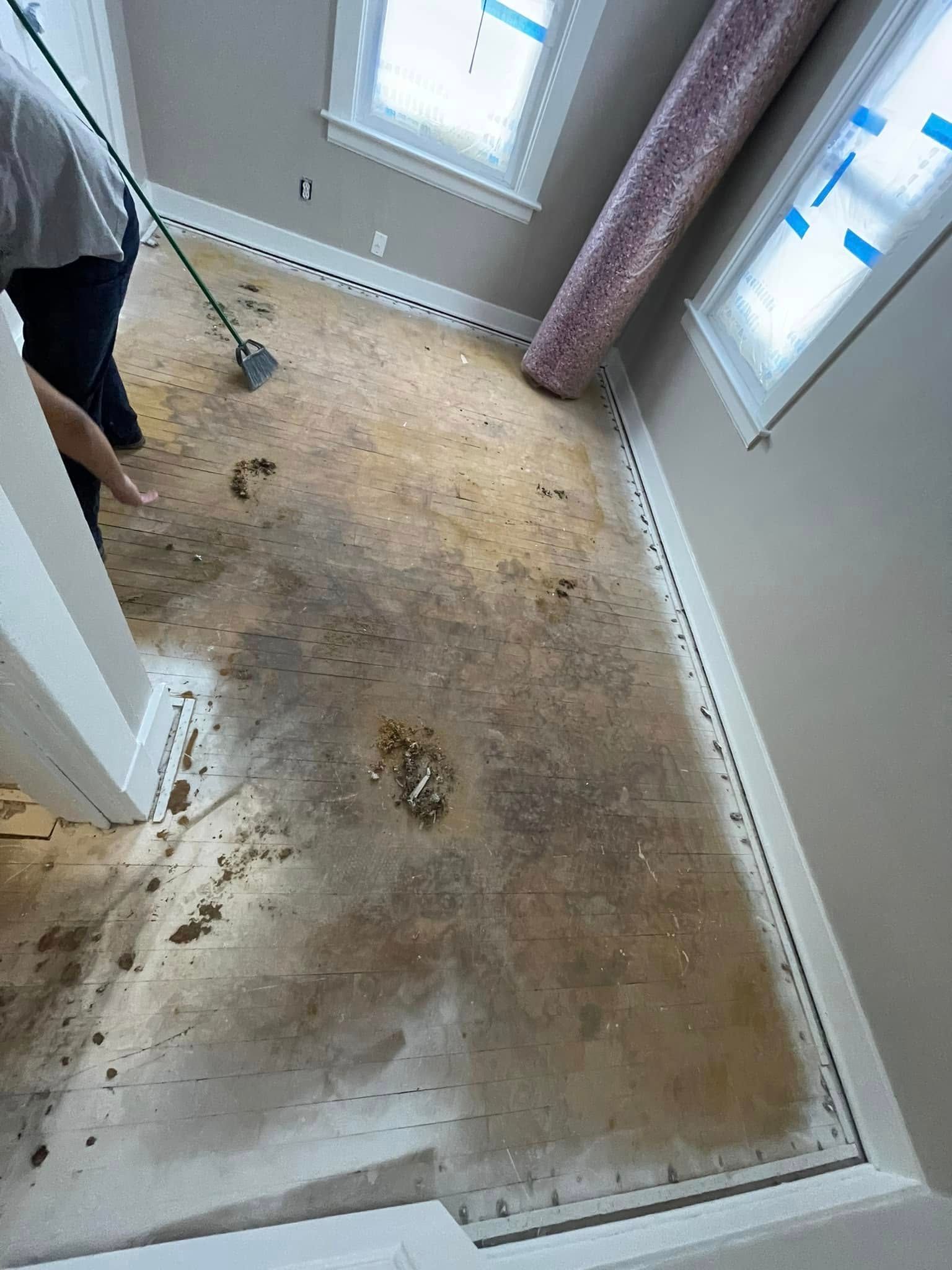 Person cleaning debris on a stained floor; two windows, a roll of carpet, and stairs are also visible.