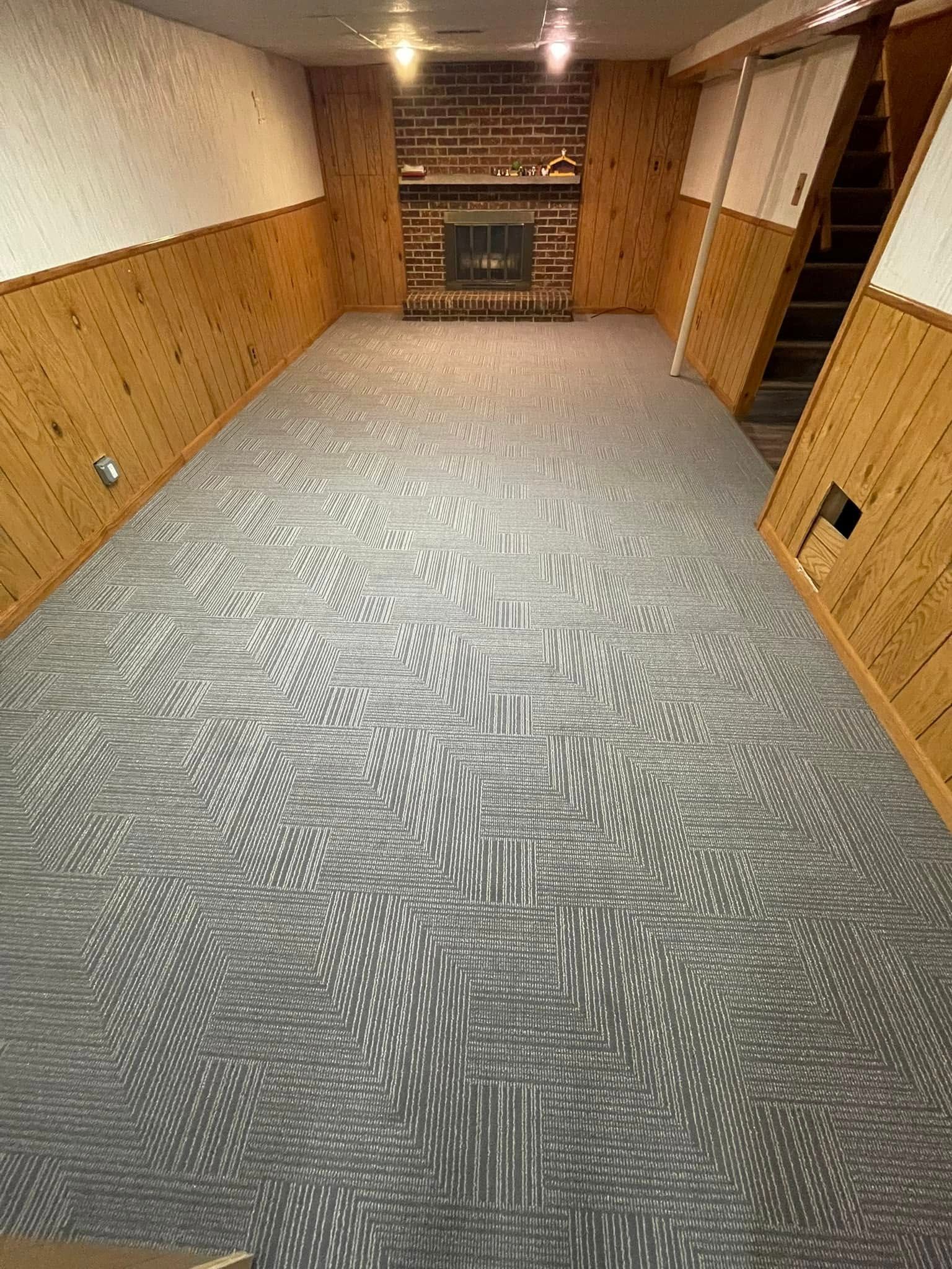 Basement with patterned gray carpet, wood-paneled walls, brick fireplace, and stairs.