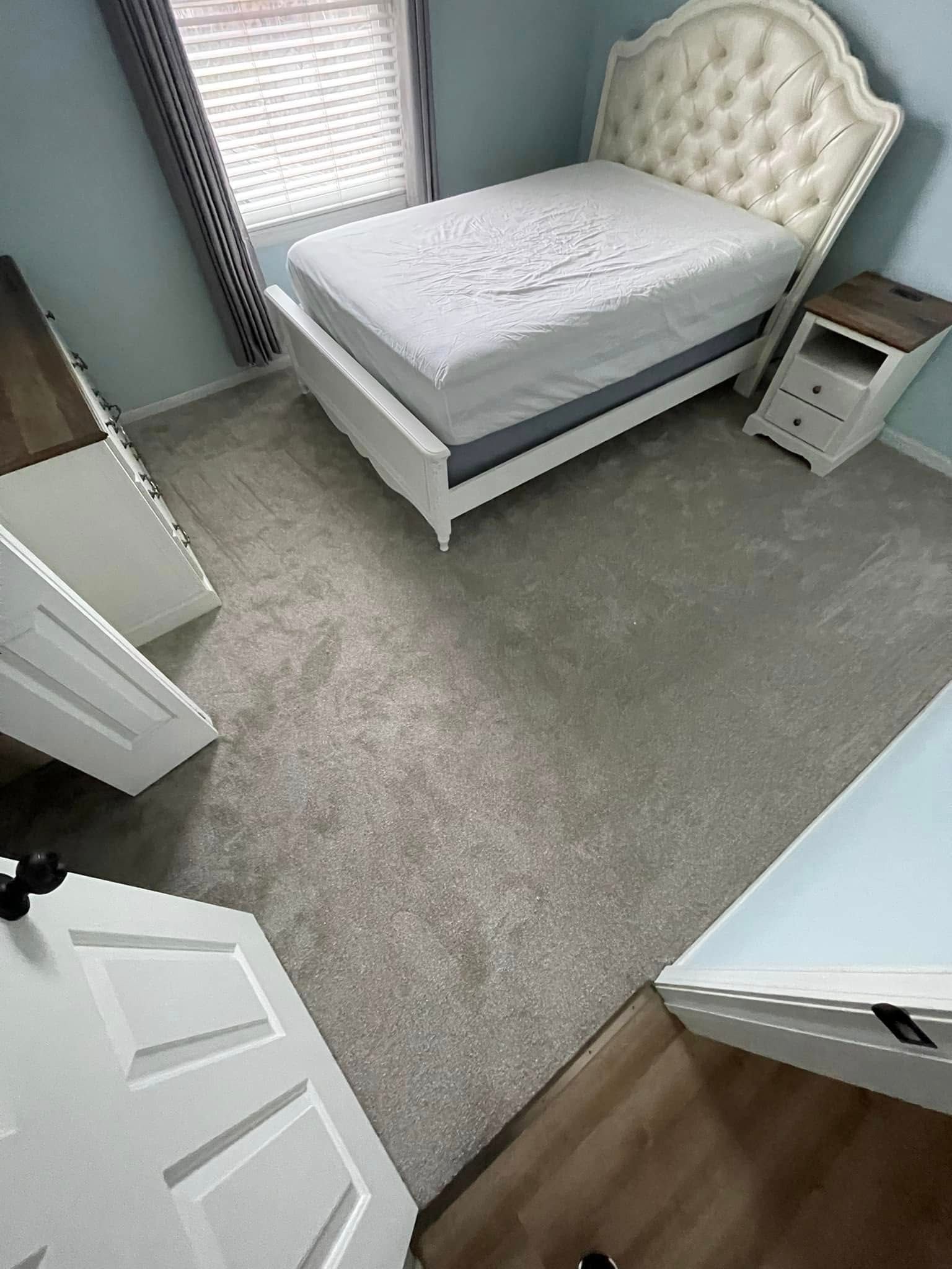 Bedroom with white bed, nightstand, dresser, door, and carpet. Window with blinds. Light blue walls.