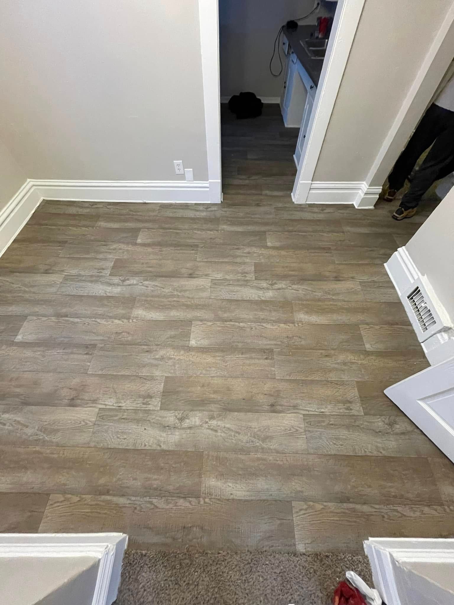 Wood-look flooring in a room with white trim and doorway. A person's legs are partially visible.