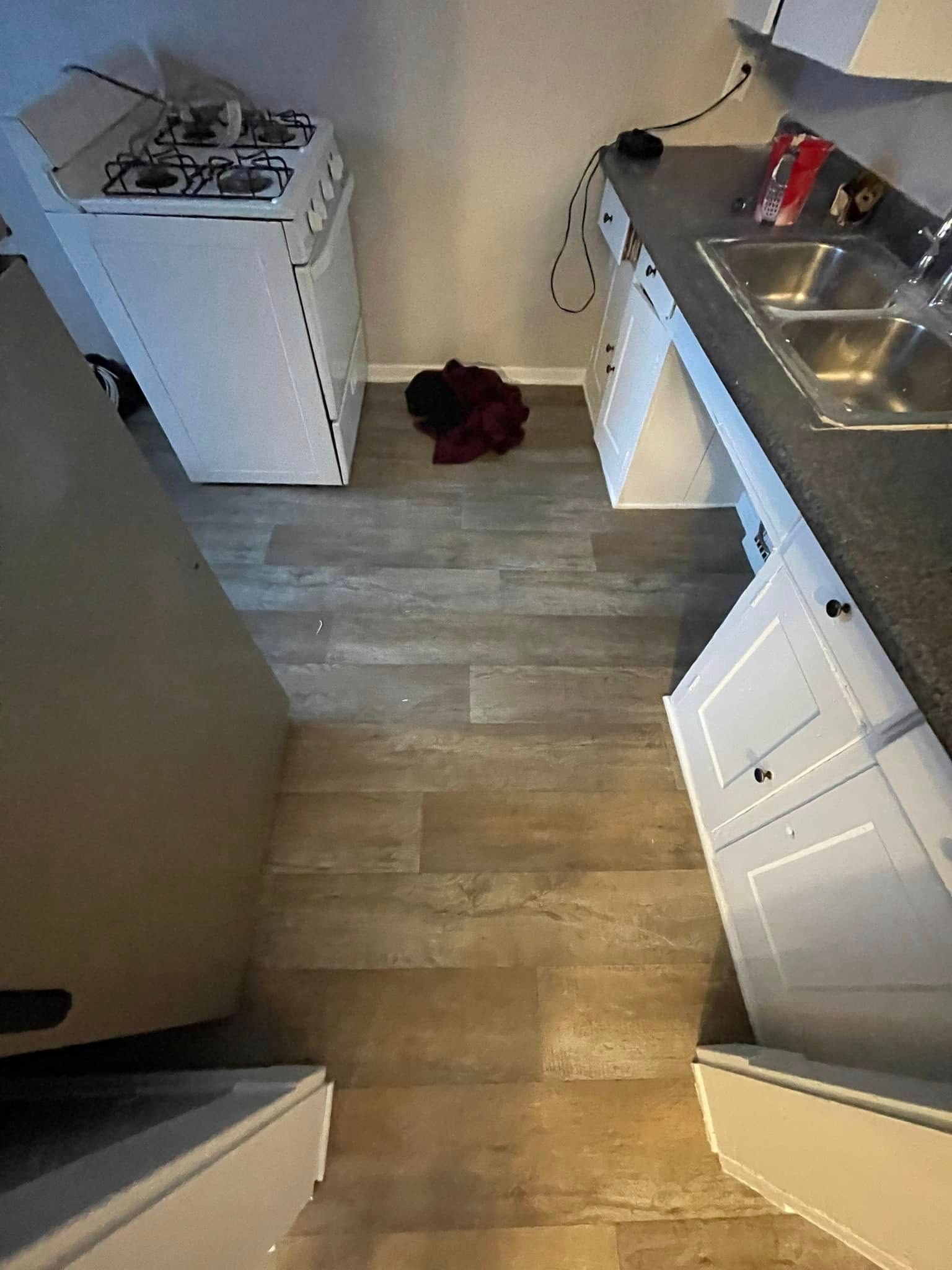 Kitchen with white cabinets, stove, sink, and wood-look flooring. A pile of clothes is in the center.