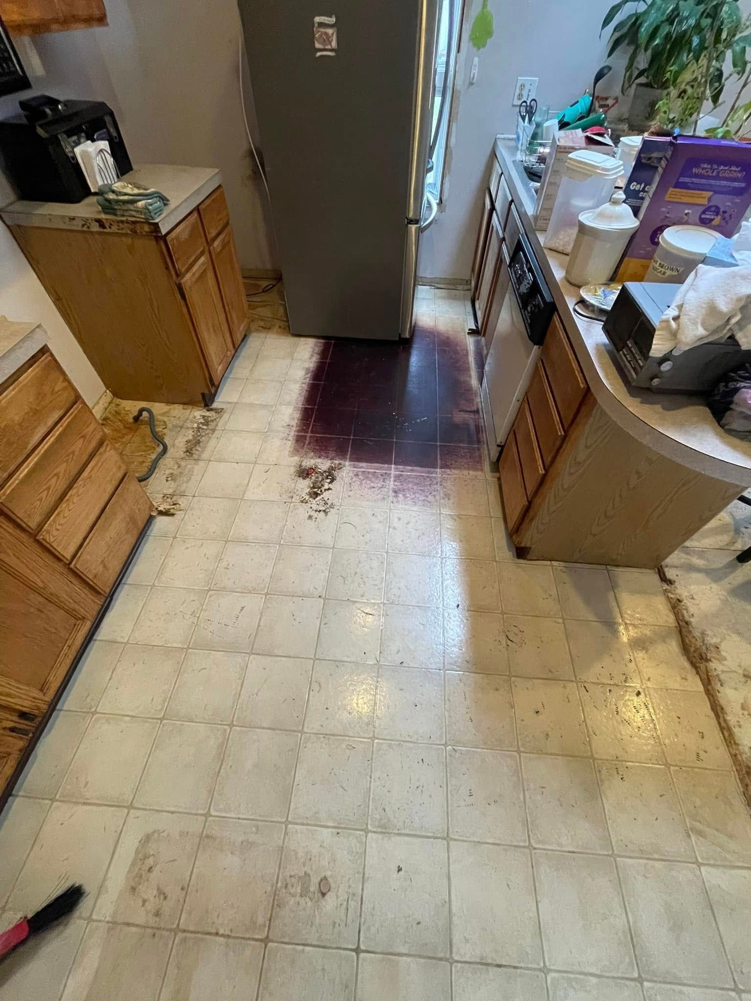Kitchen floor with stained tiles, cabinets, refrigerator, and countertop clutter.