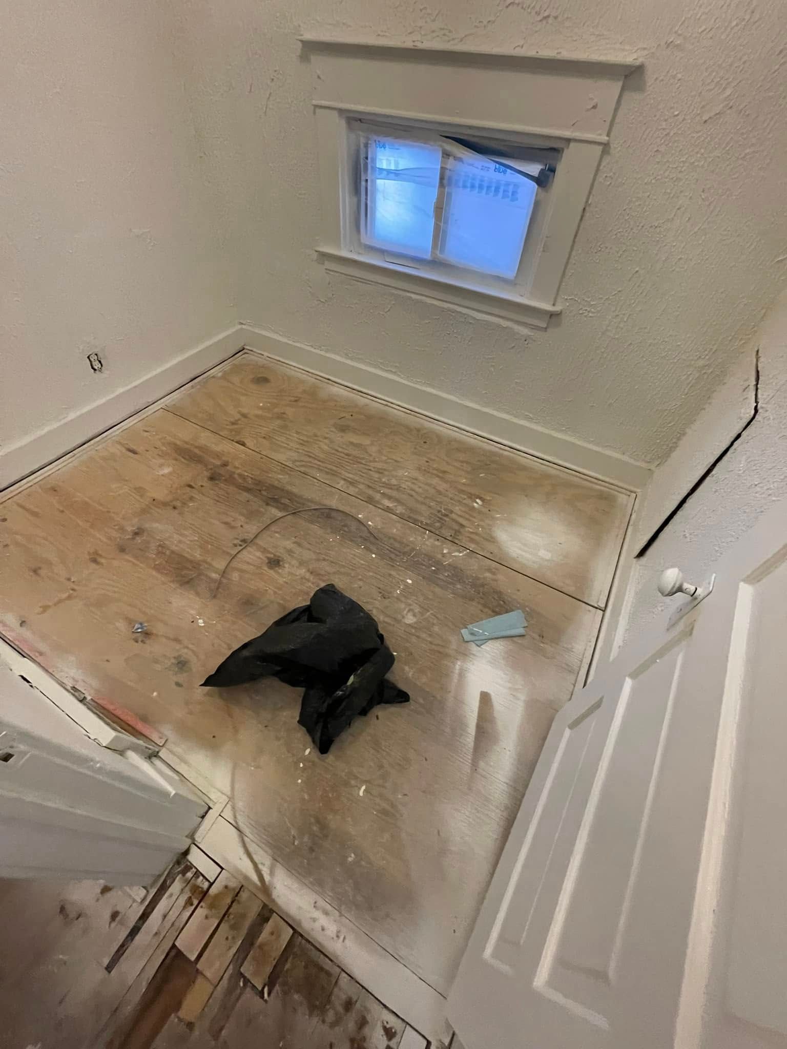 Room under construction; wooden floor, small window, black trash bag, white walls, and doorway.
