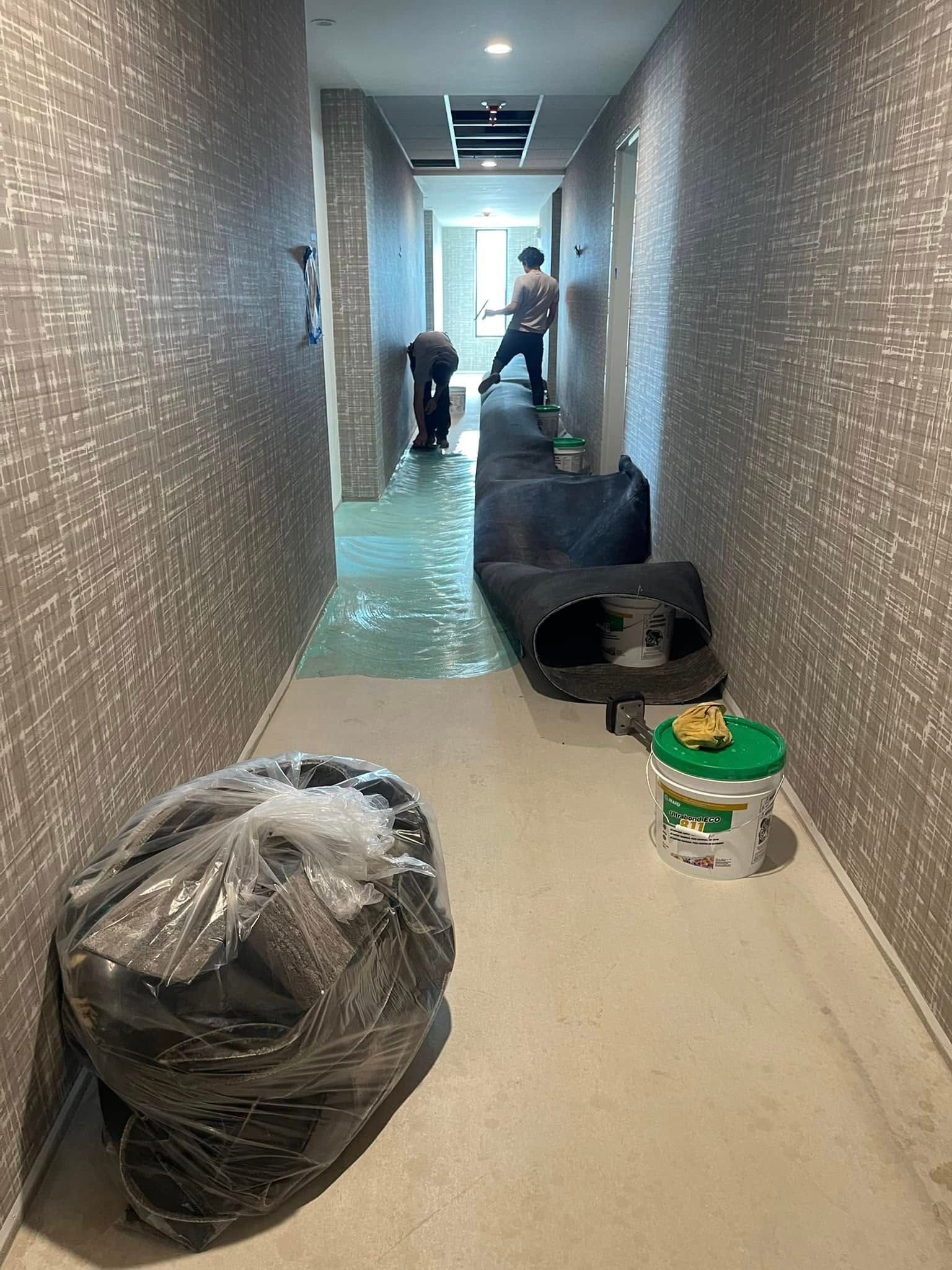 Workers in hallway with wet floor, debris, and garbage bag. Walls are tiled.