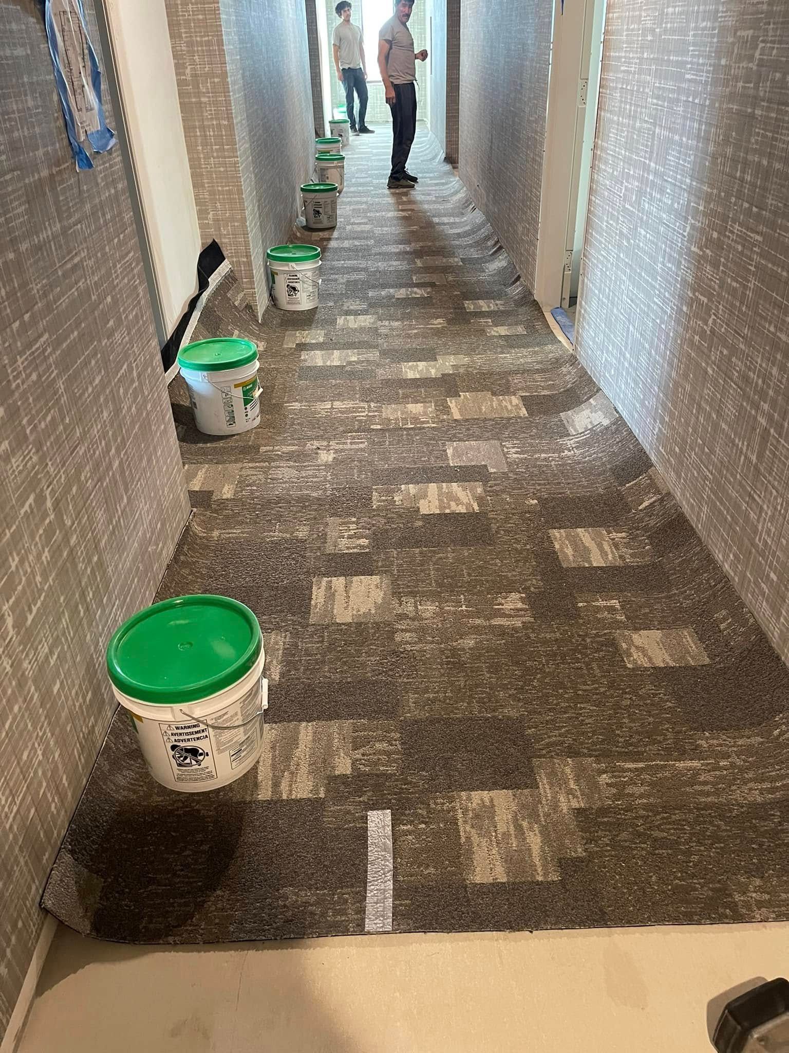 Hallway with carpet installation in progress; workers in the distance, buckets, and partially laid carpet visible.