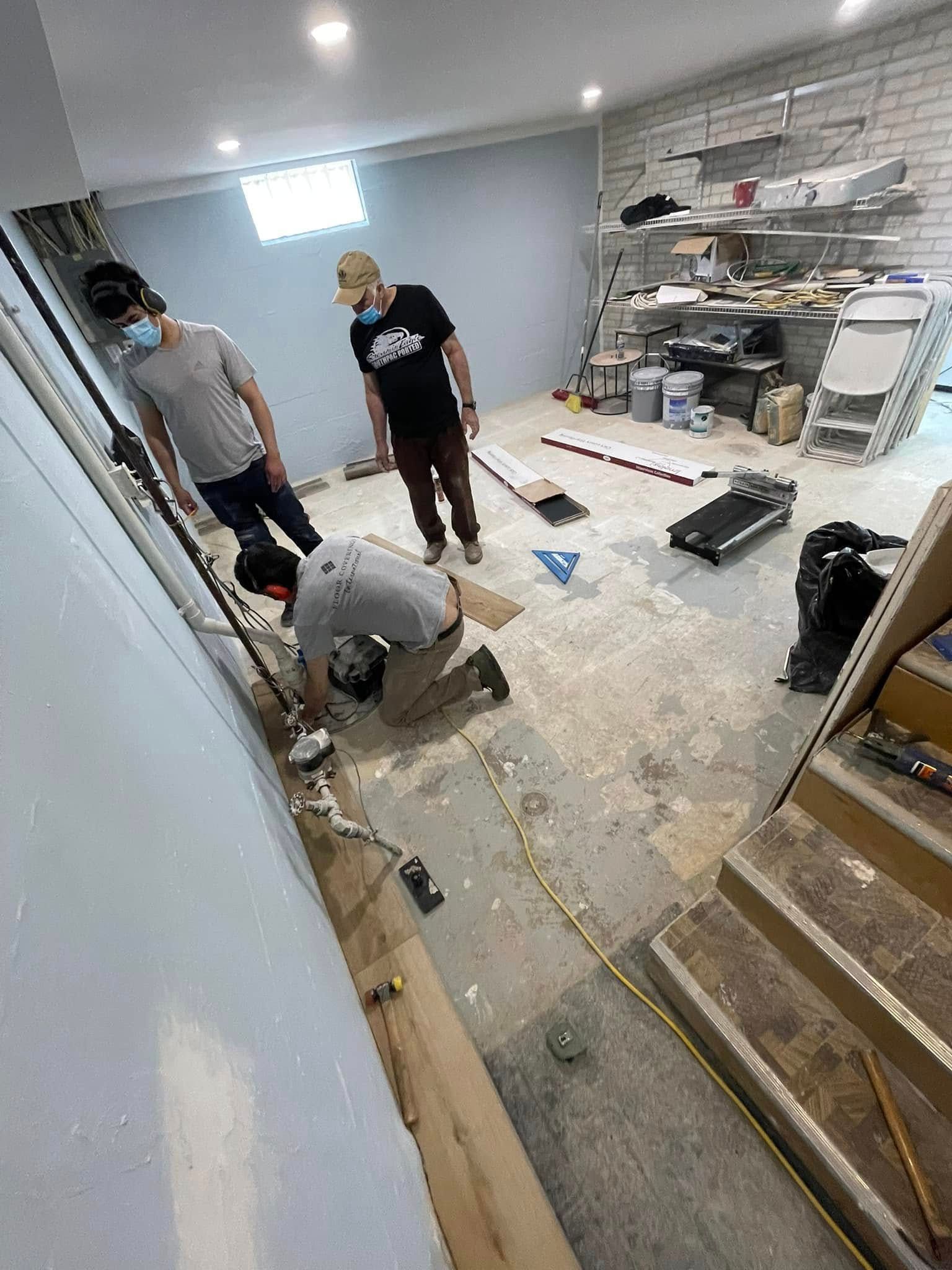 Three people in a basement, one kneeling, working on flooring. Gray walls and exposed concrete.
