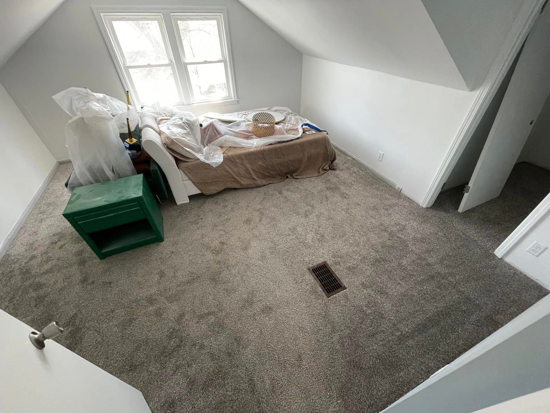 Bedroom with light-colored walls and carpet. A bed is covered in plastic. A green nightstand sits next to the bed.
