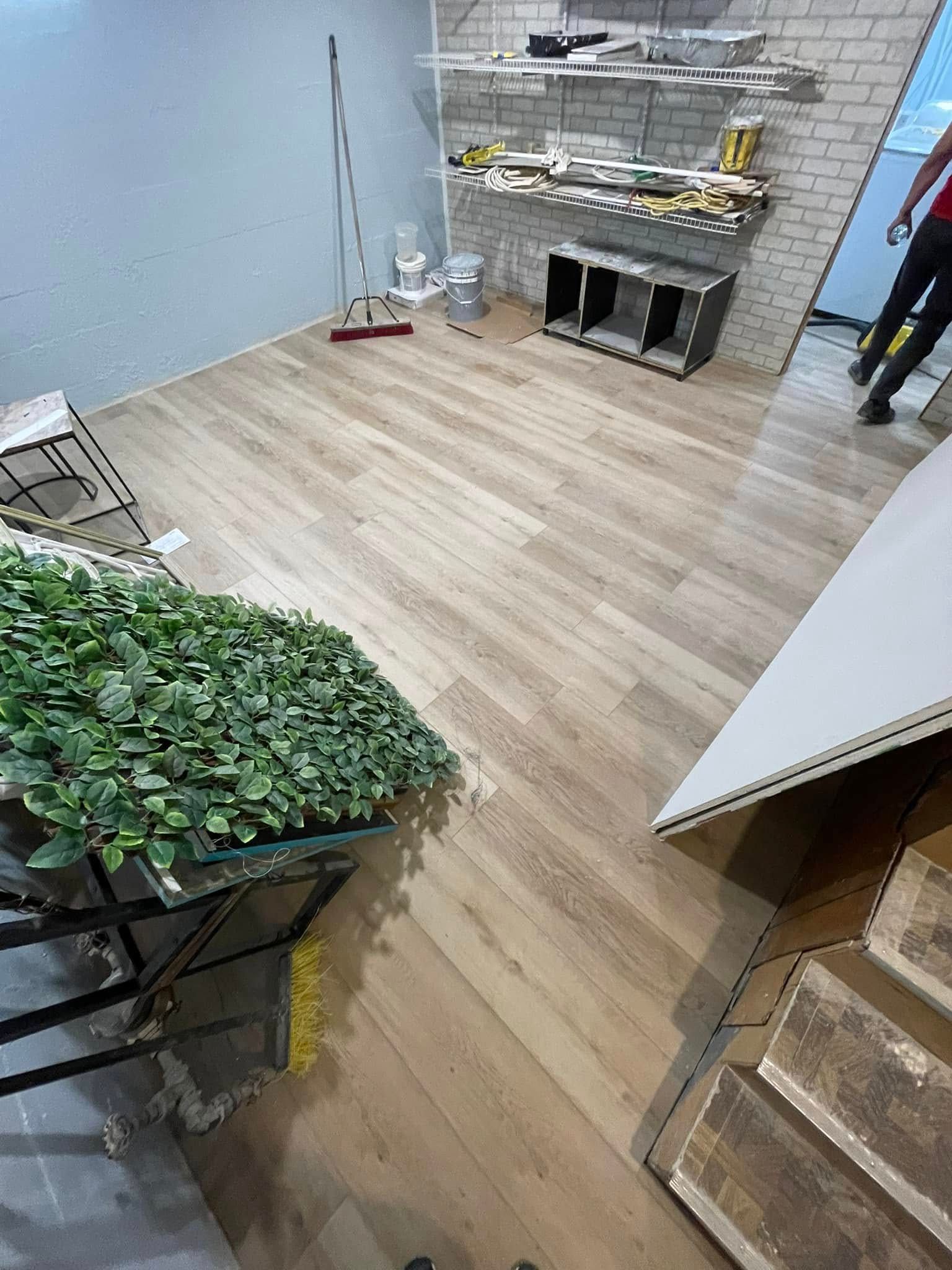 Light-wood floors in a room under renovation. Artificial greenery, exposed brick, and a person in the background.