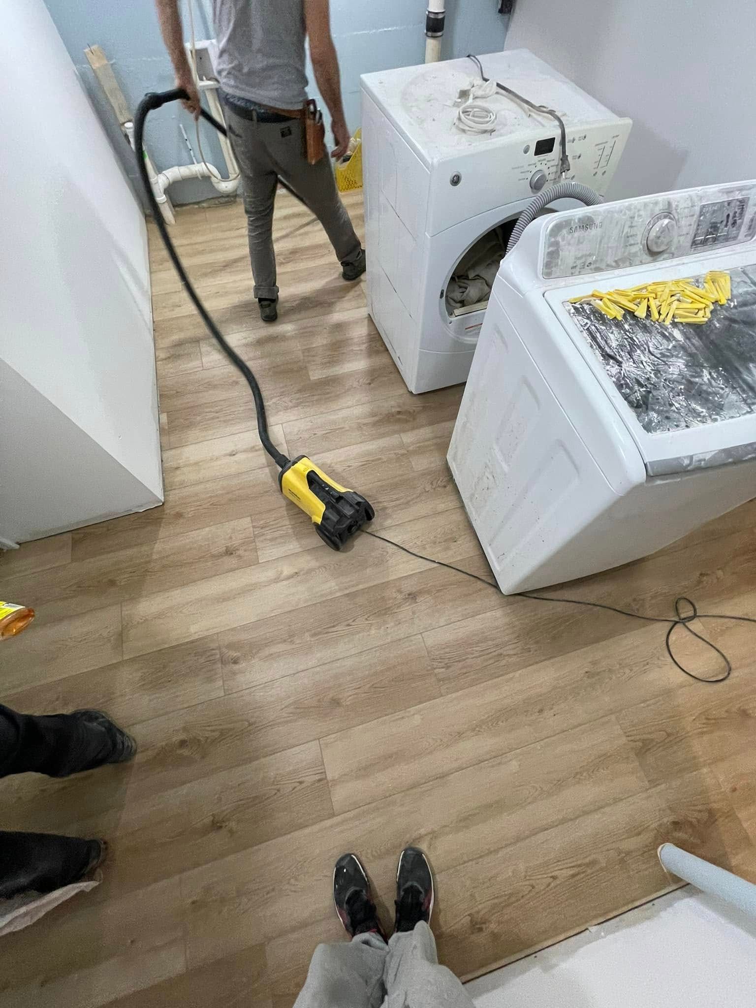 Person vacuums floor near washing machine and dryer in a room with light wood floors.