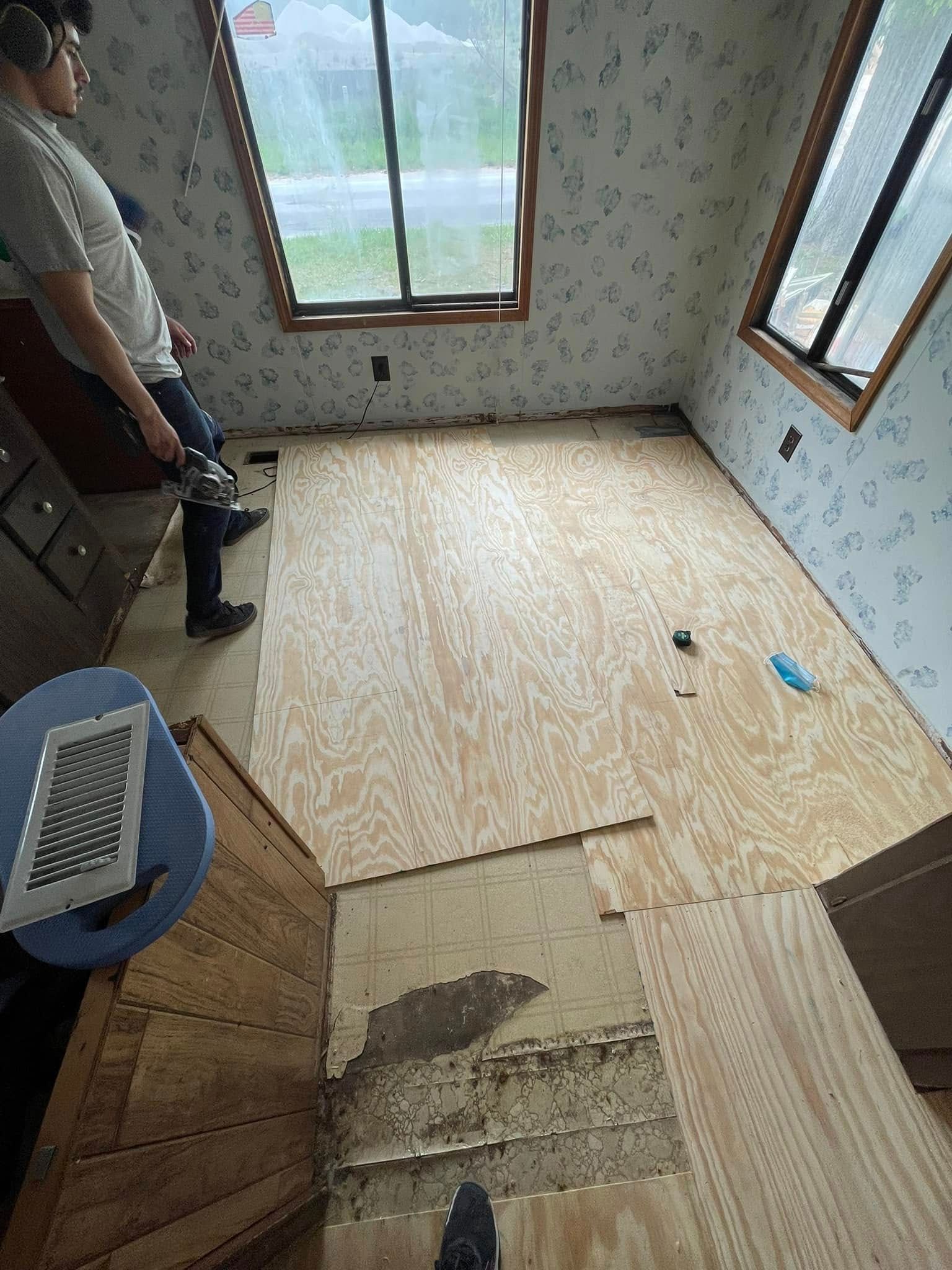 Person using tool to work on plywood flooring in a room with two windows.