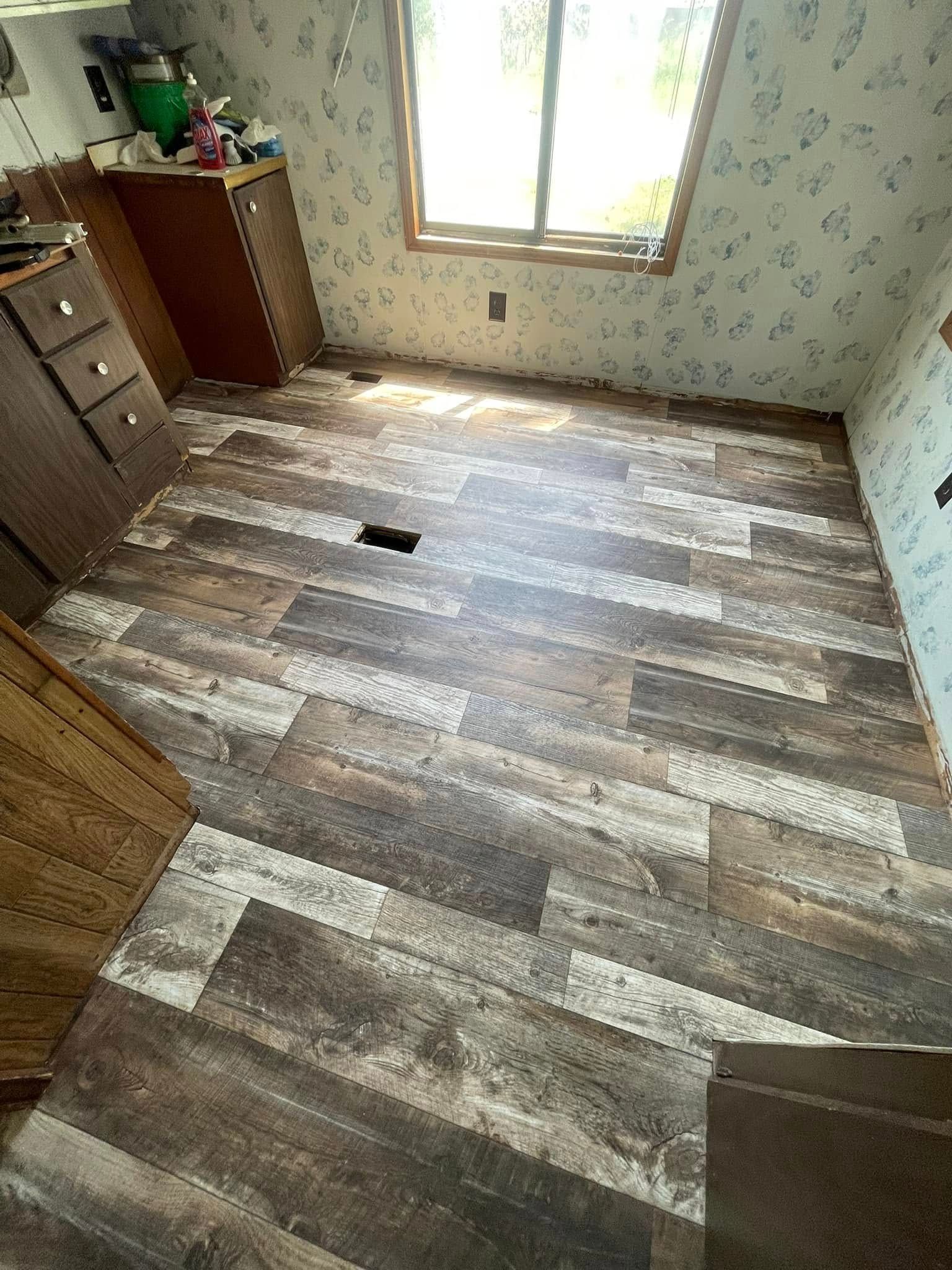 Room with new wood-look flooring installed. Window at top. Cabinets along left wall. Wallpapered walls.