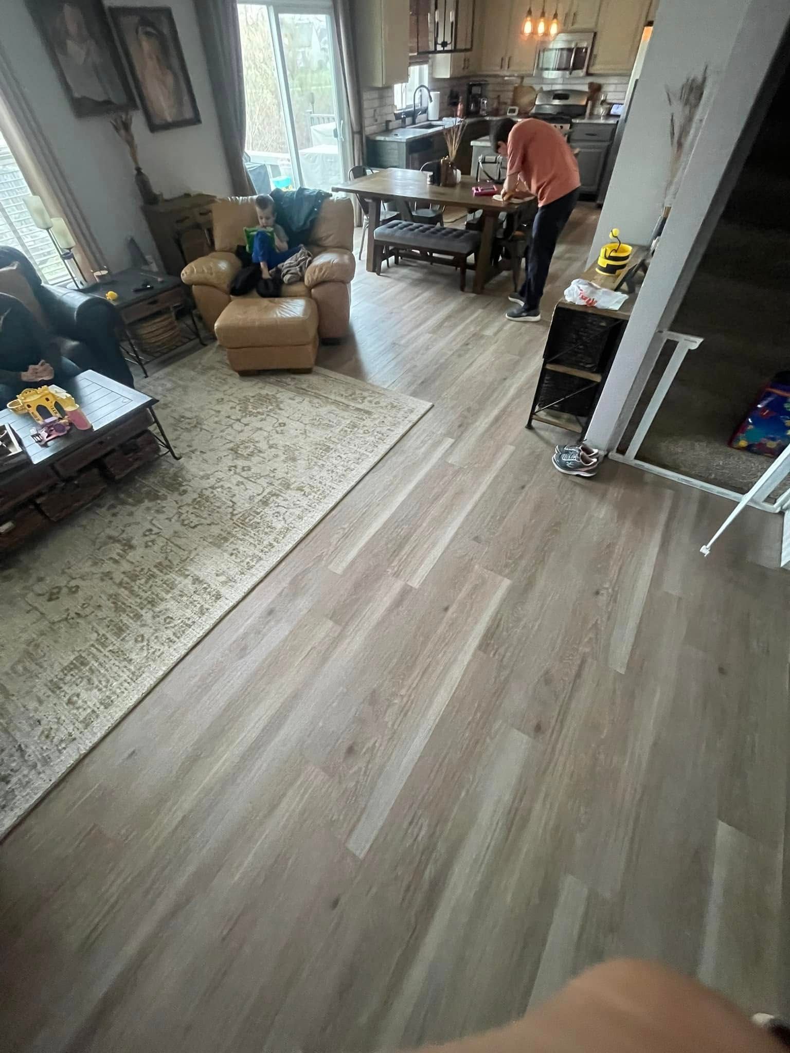 Light wood-look flooring extends through a living area. A person stands near a table in the background. A rug is on the floor.