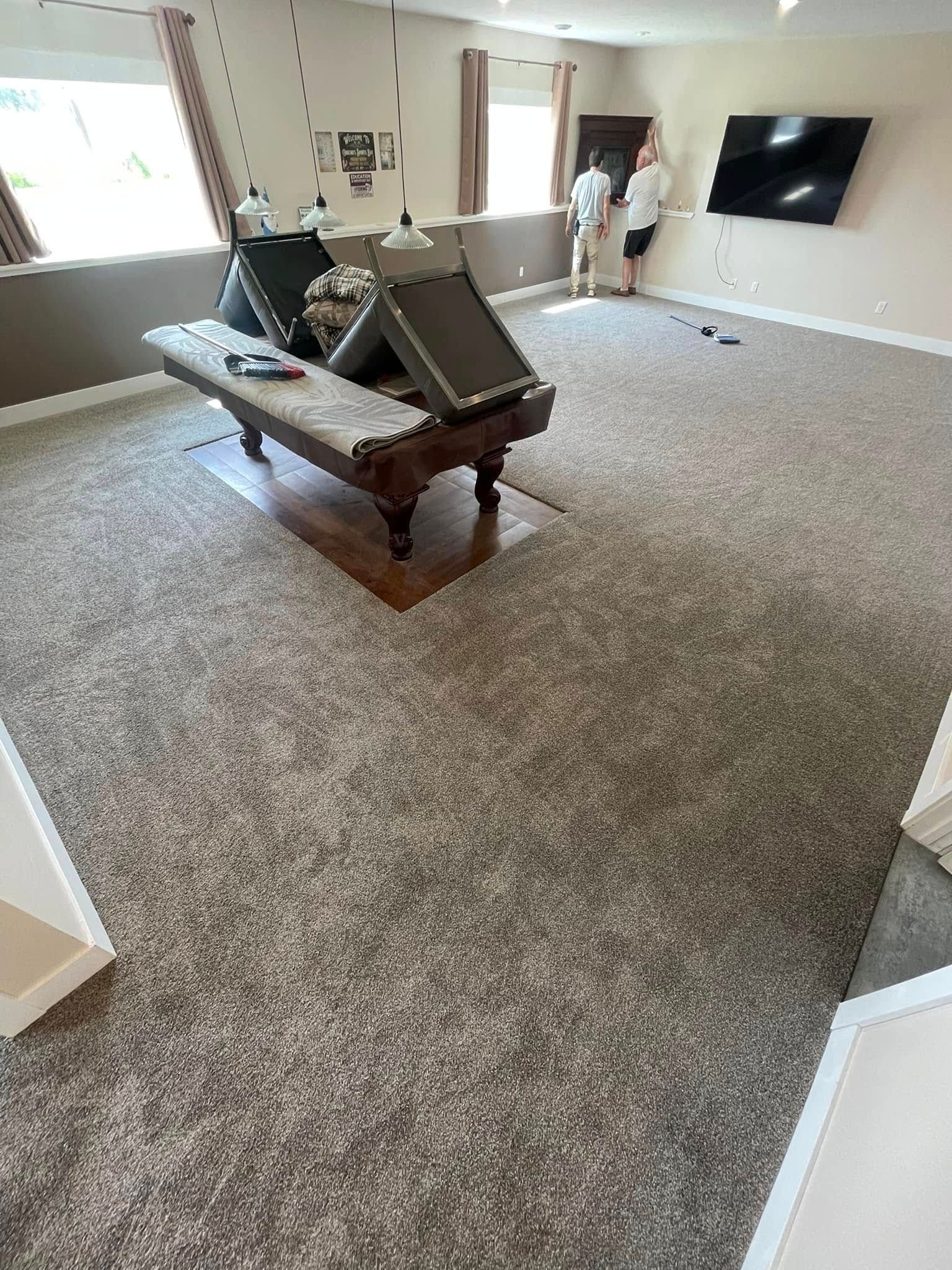 A large room with a pool table and two people near a TV on the wall. Brown carpet.