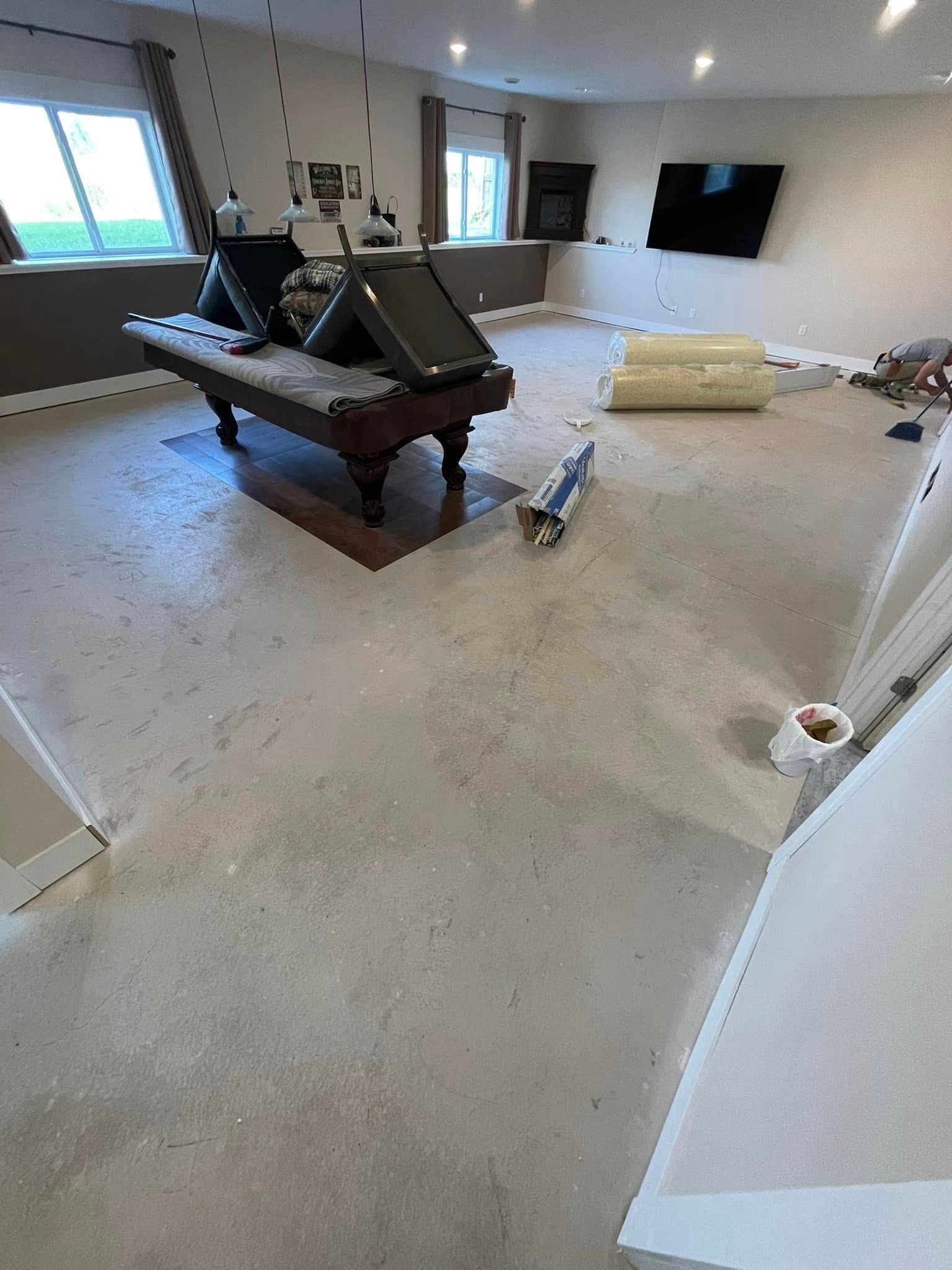 A basement room with a pool table, rolled-up carpet, and construction materials on a concrete floor.