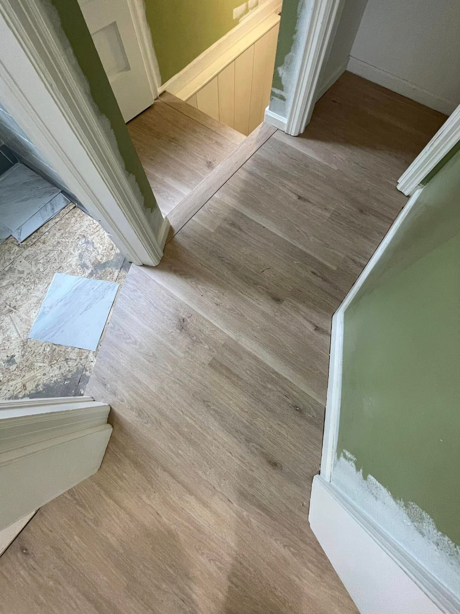 Wood flooring in a hallway with doorways. Light brown planks, light green walls, white trim.
