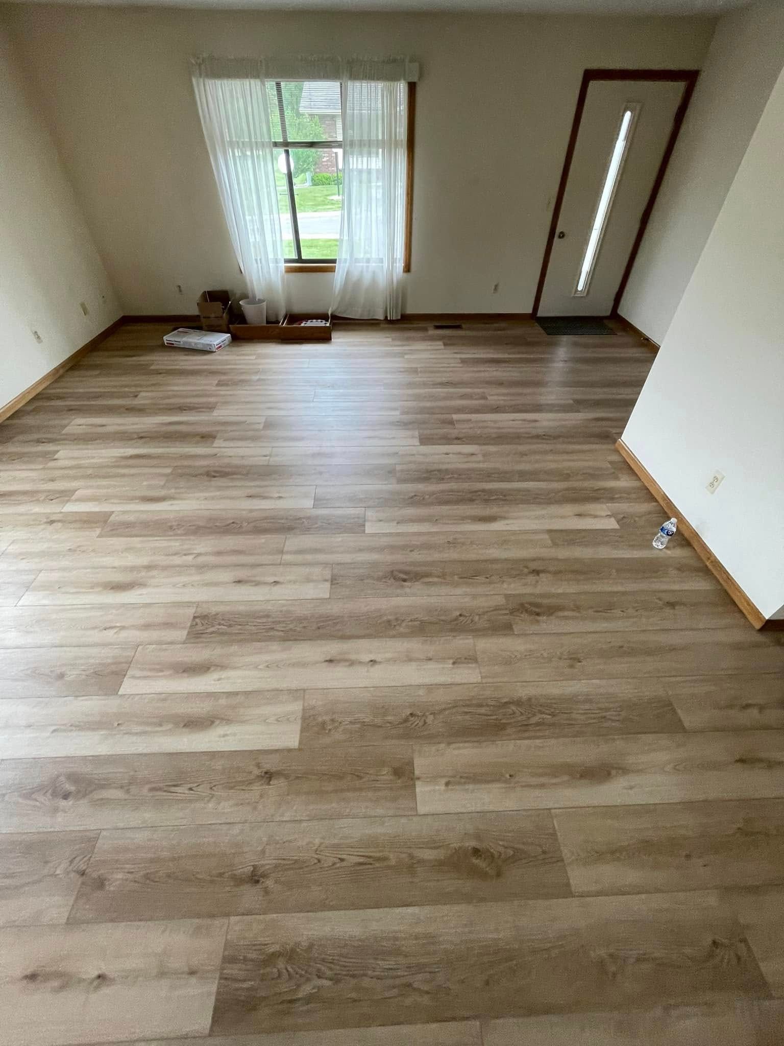 Empty room with light-brown wood-look flooring, white walls, window with sheer curtains, and a door.