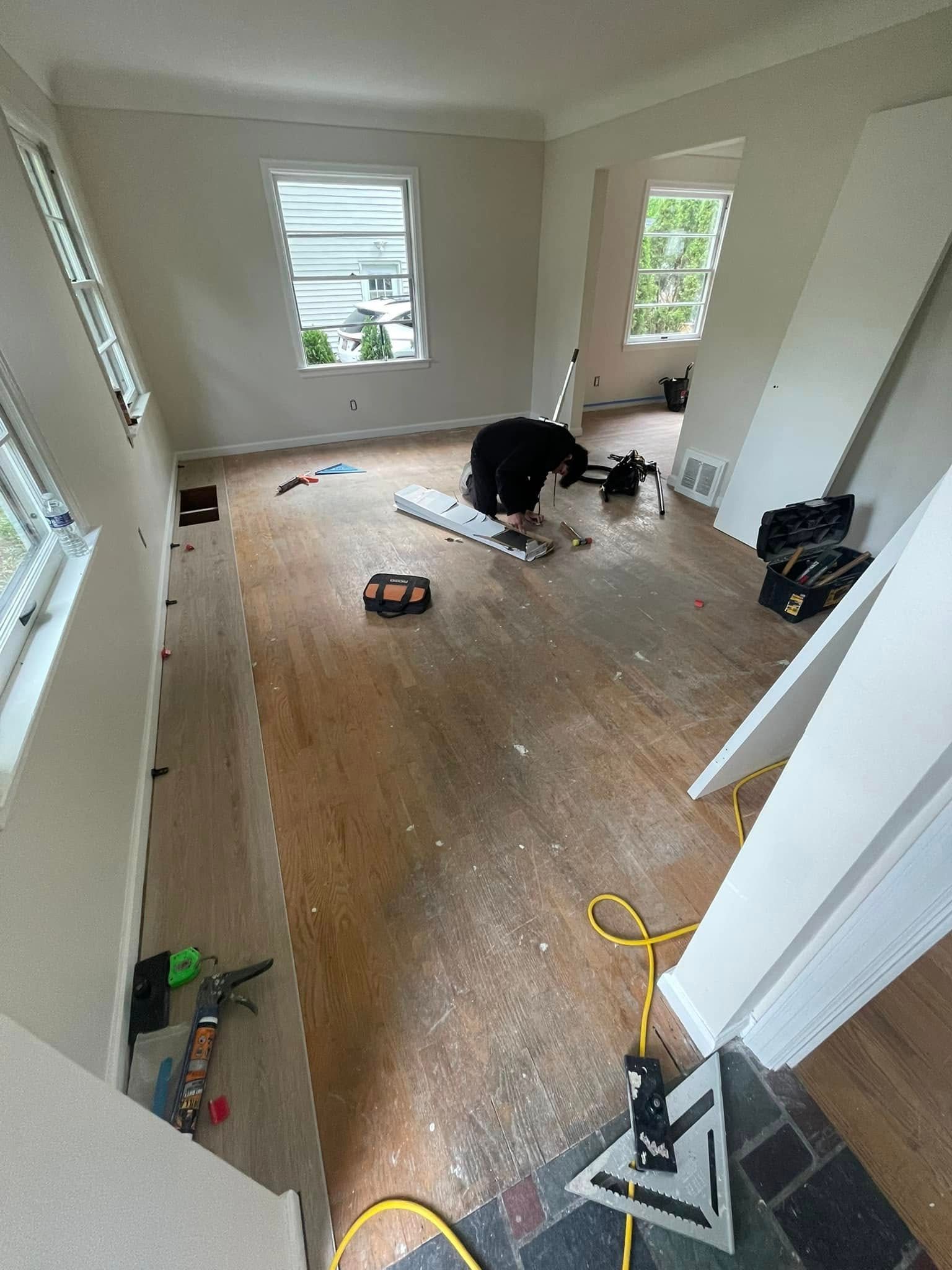 Person installing wooden flooring in a room with white walls and windows. Tools and materials are scattered.