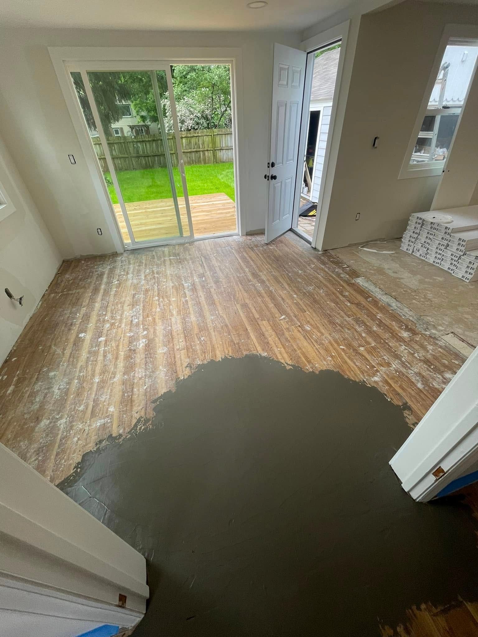Interior room with hardwood floor, partially covered in gray material, leading to a backyard.
