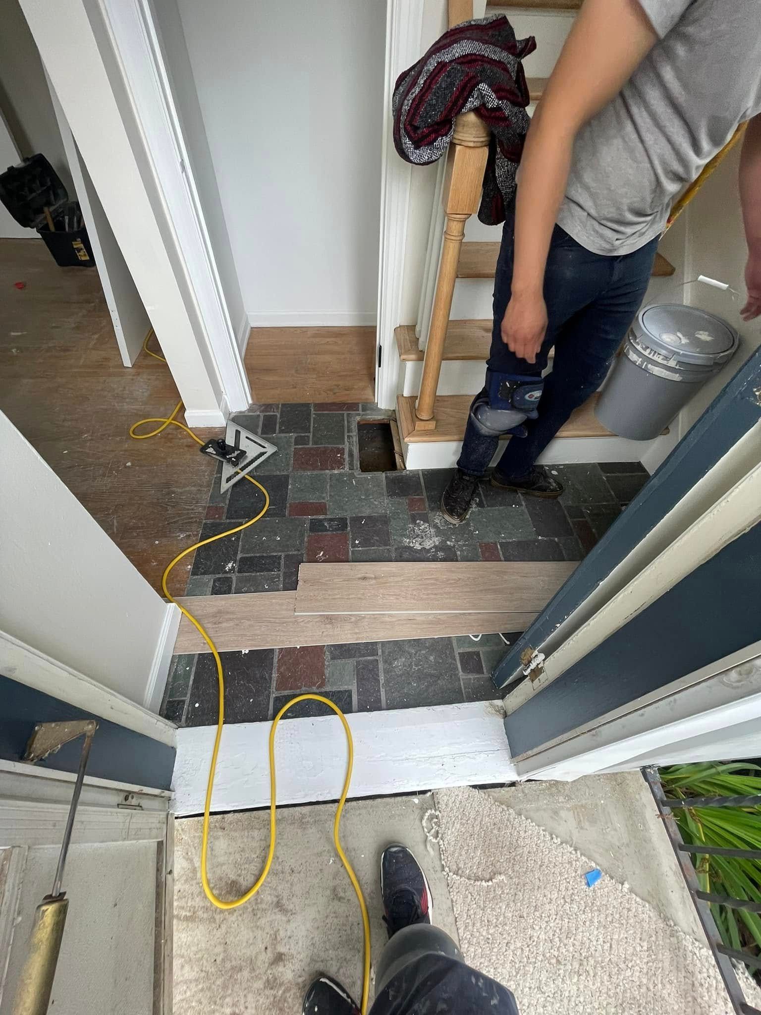 Person standing near stairs and doorway, renovations in progress; flooring removed, tools and debris visible.
