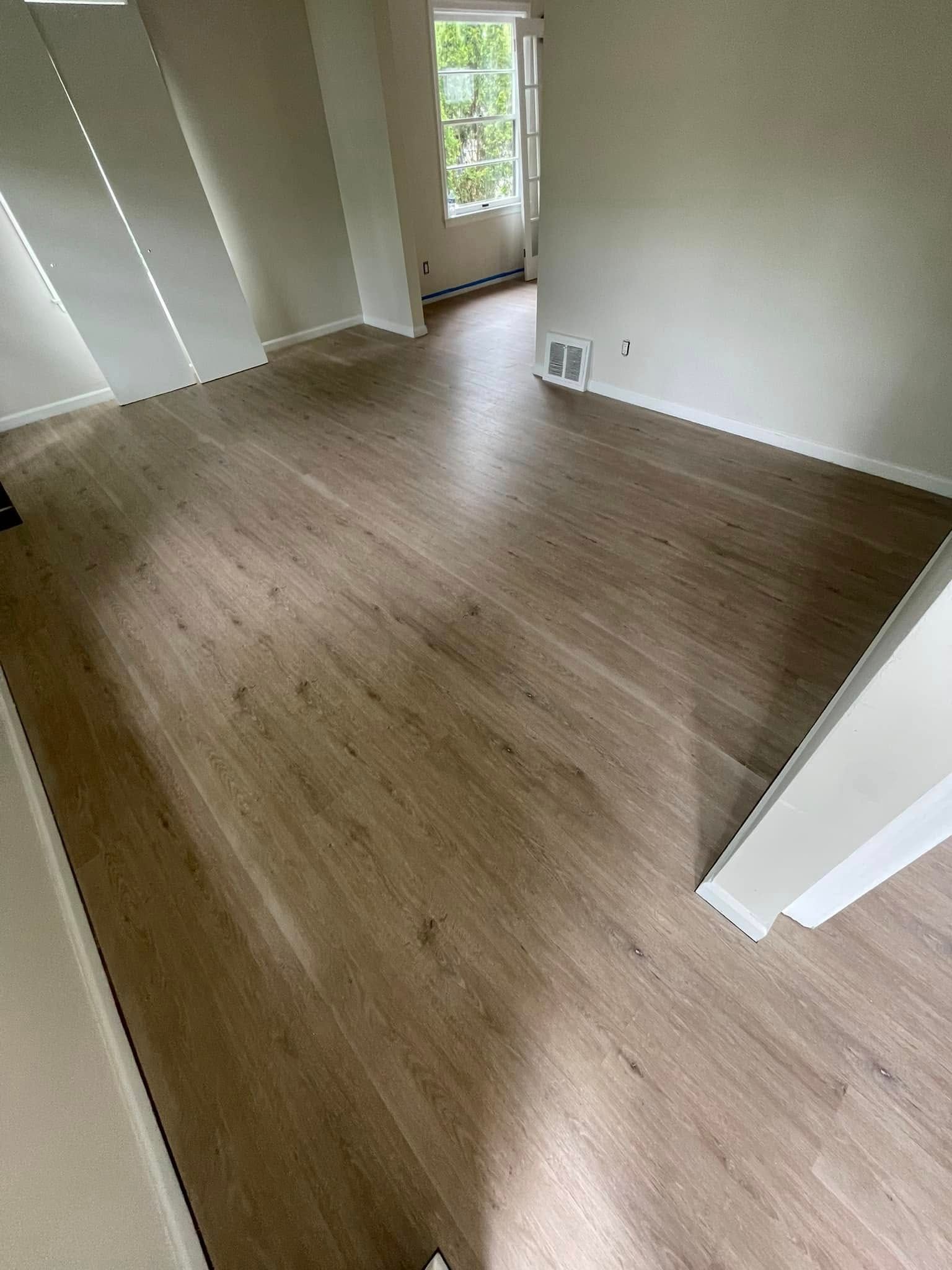 Empty room with light wood-look flooring and white walls. A doorway leads to a window with natural light.