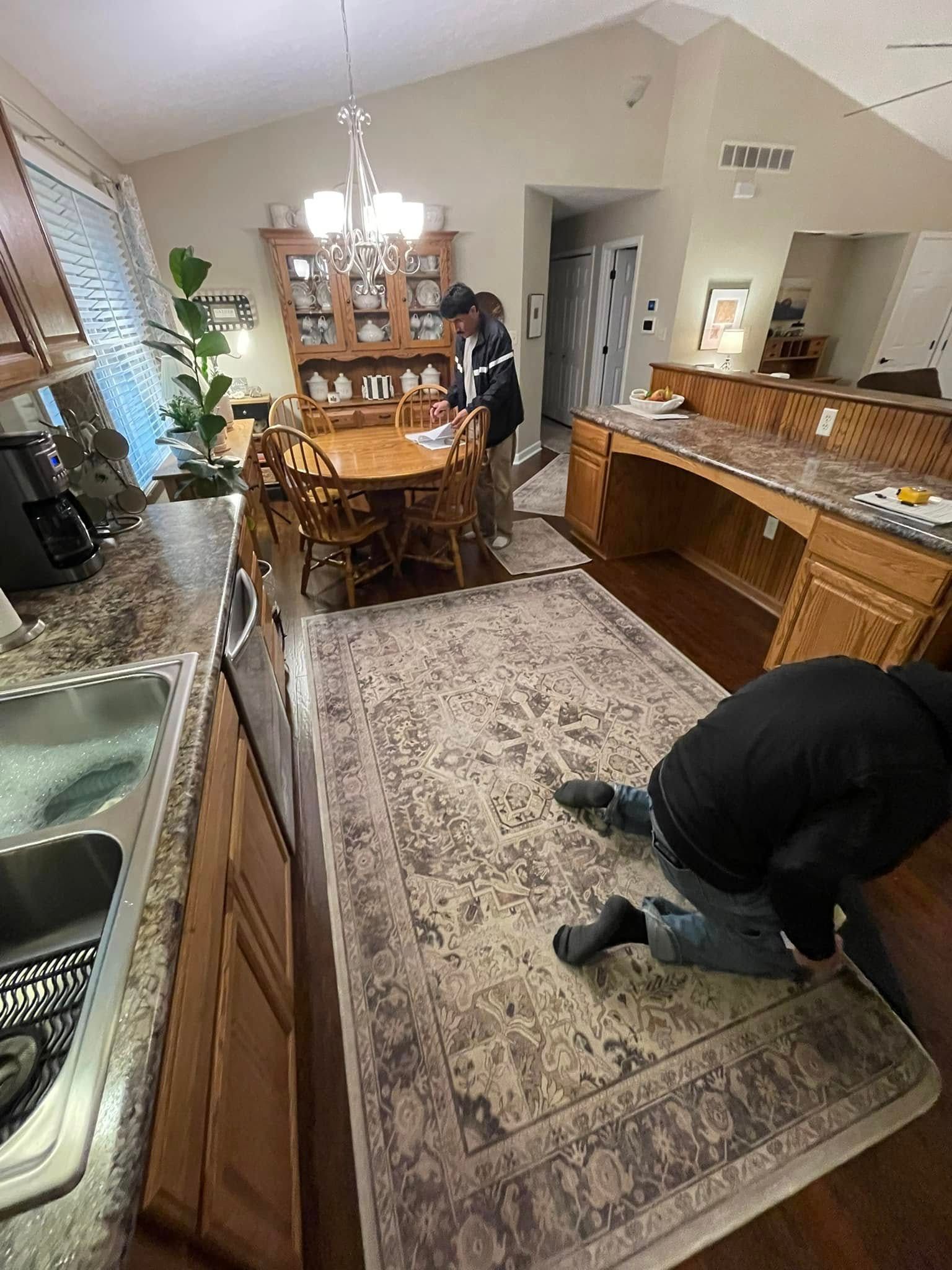 Person kneeling, laying rug in dining room. Kitchen counters, table, chairs, and cabinets present.