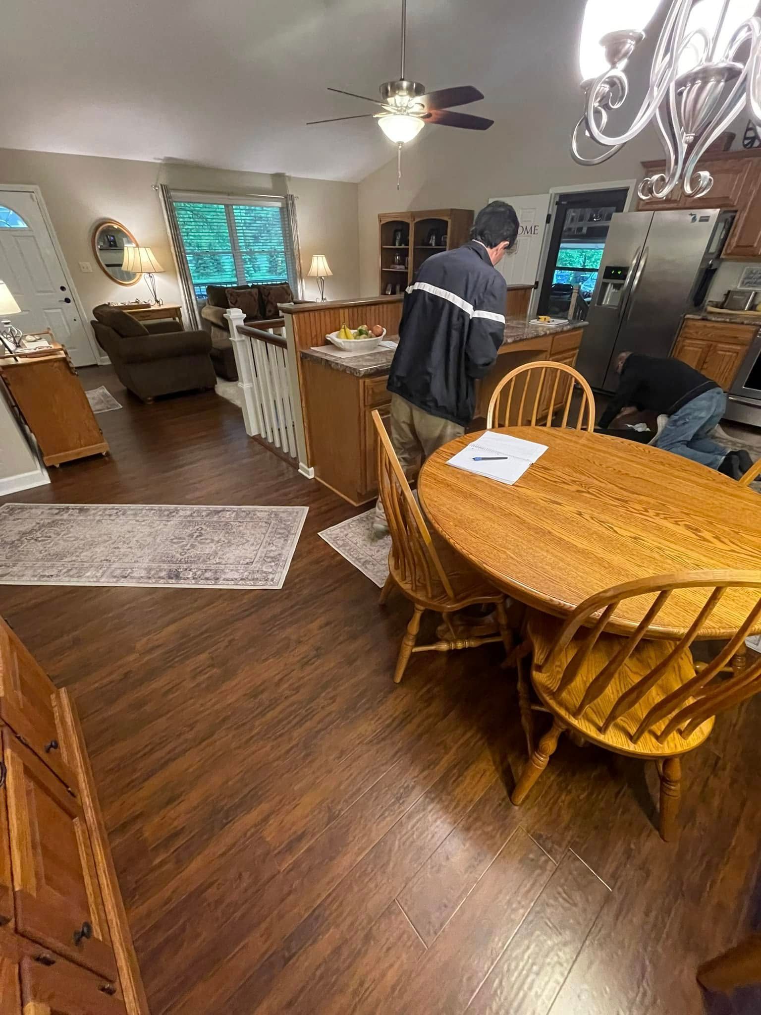 Two people in a kitchen; one by a refrigerator, the other near a table.  Wooden floors, cabinets, and a ceiling fan are visible.