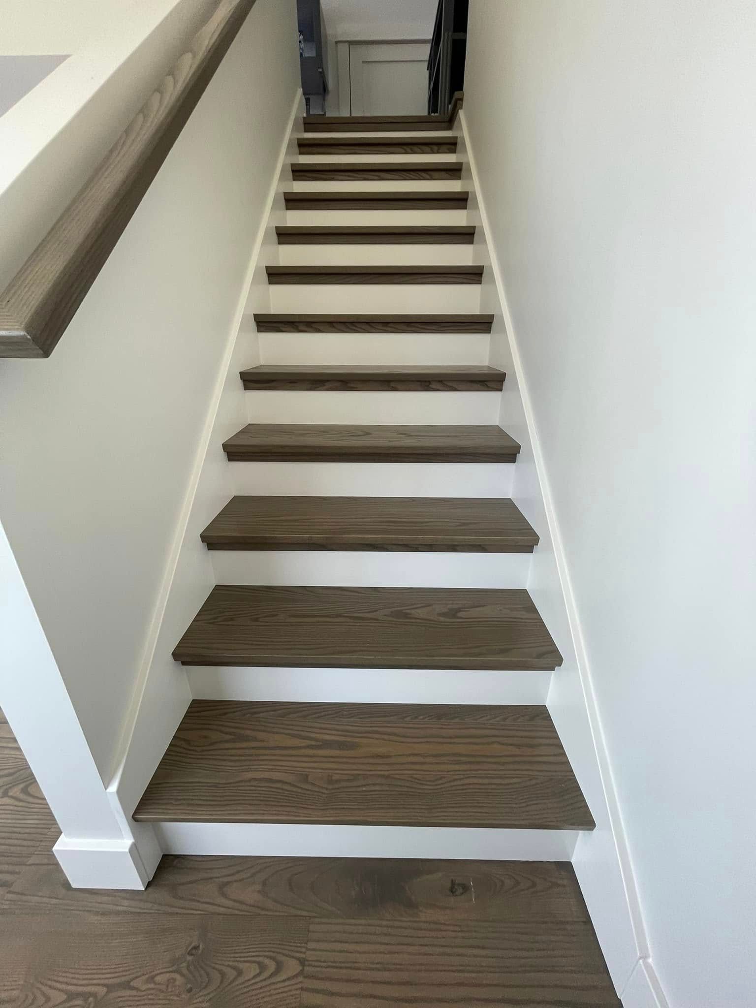 Wooden staircase with white risers and handrail, leading upwards, set against white walls.