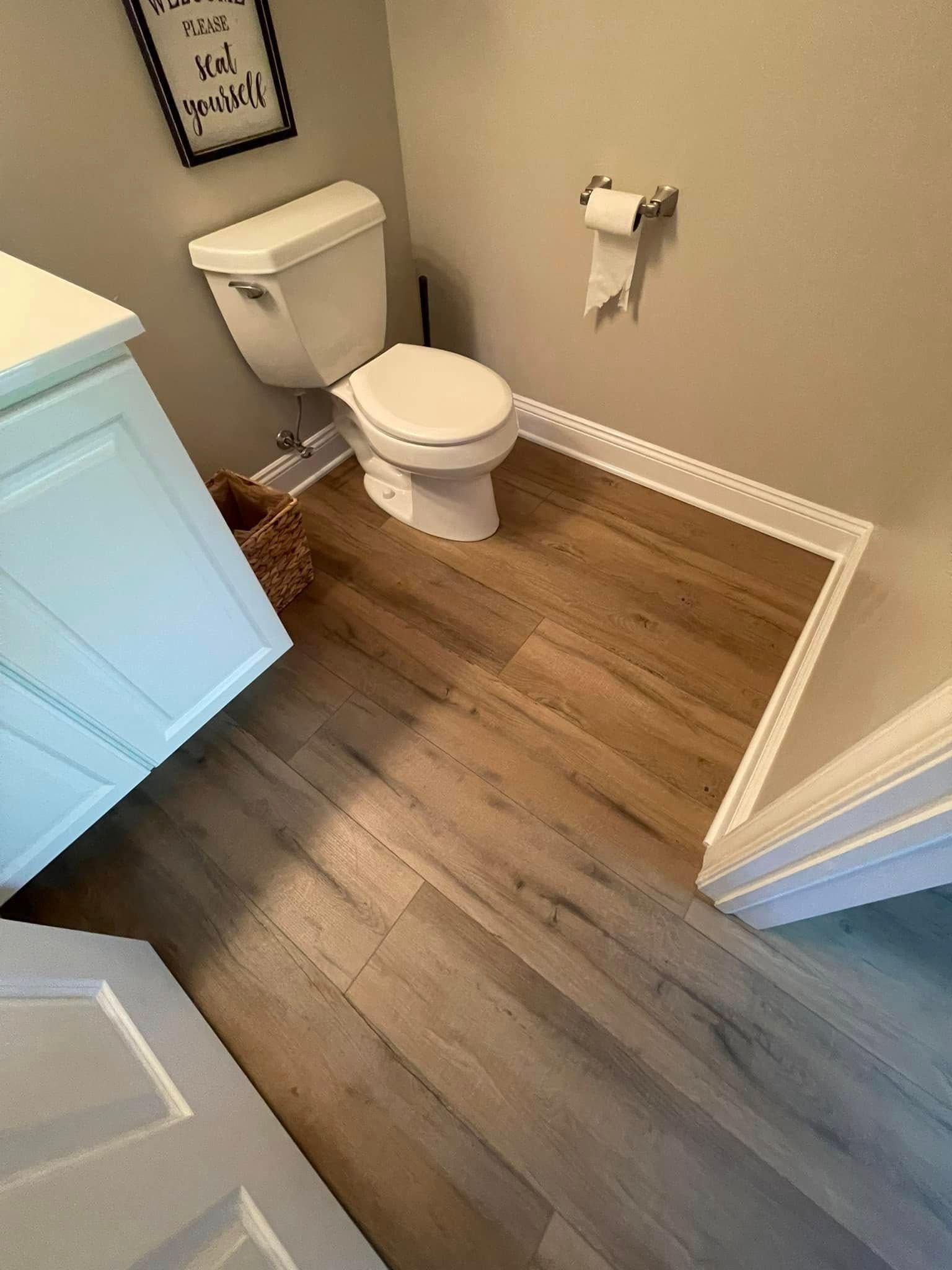Bathroom with a white toilet, cabinet, and wood-look flooring. Beige walls with a framed sign and toilet paper holder.