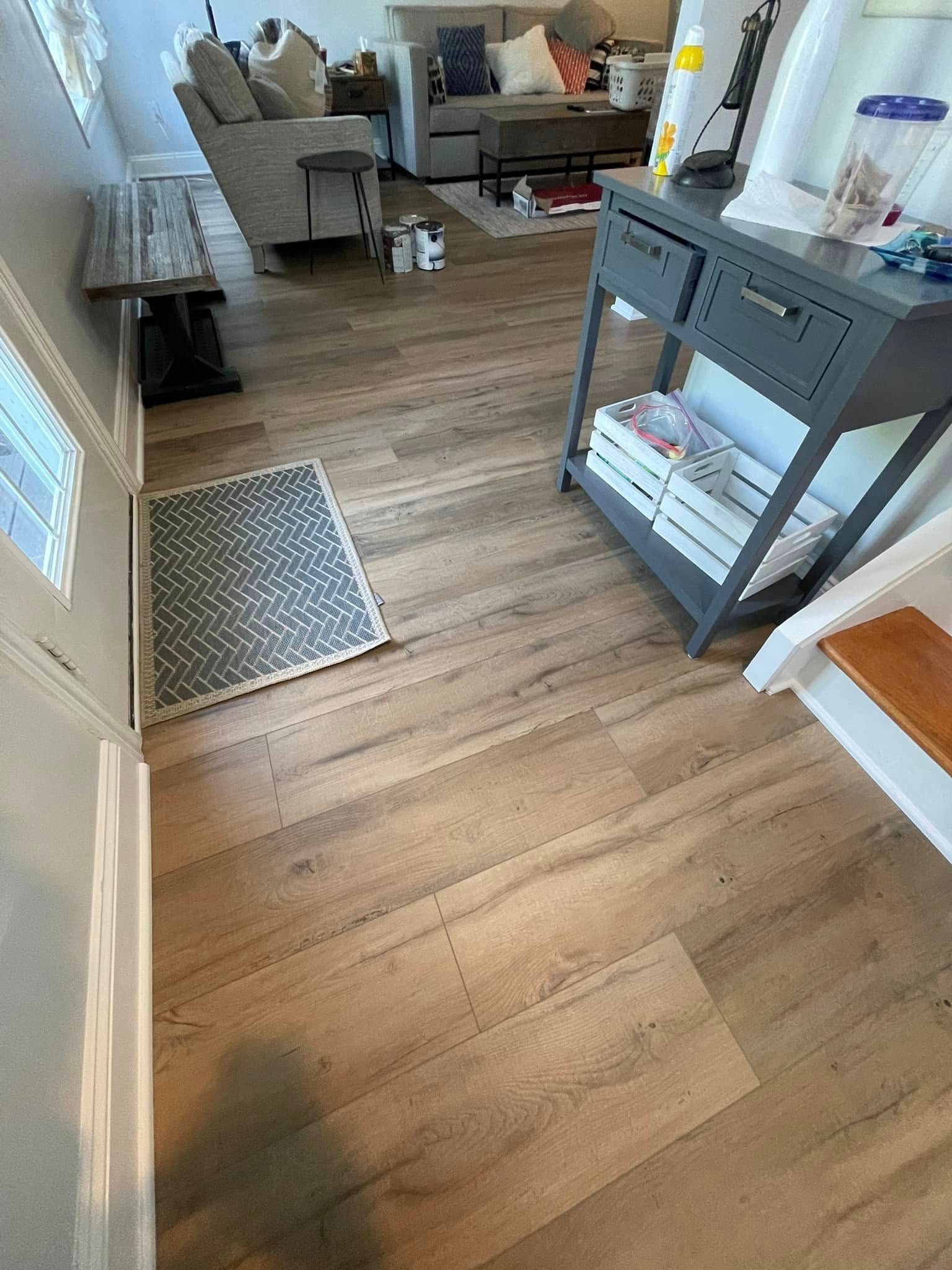 Light brown wood floor in a living room, entryway, and hallway. A gray bench, rug, and table are visible.