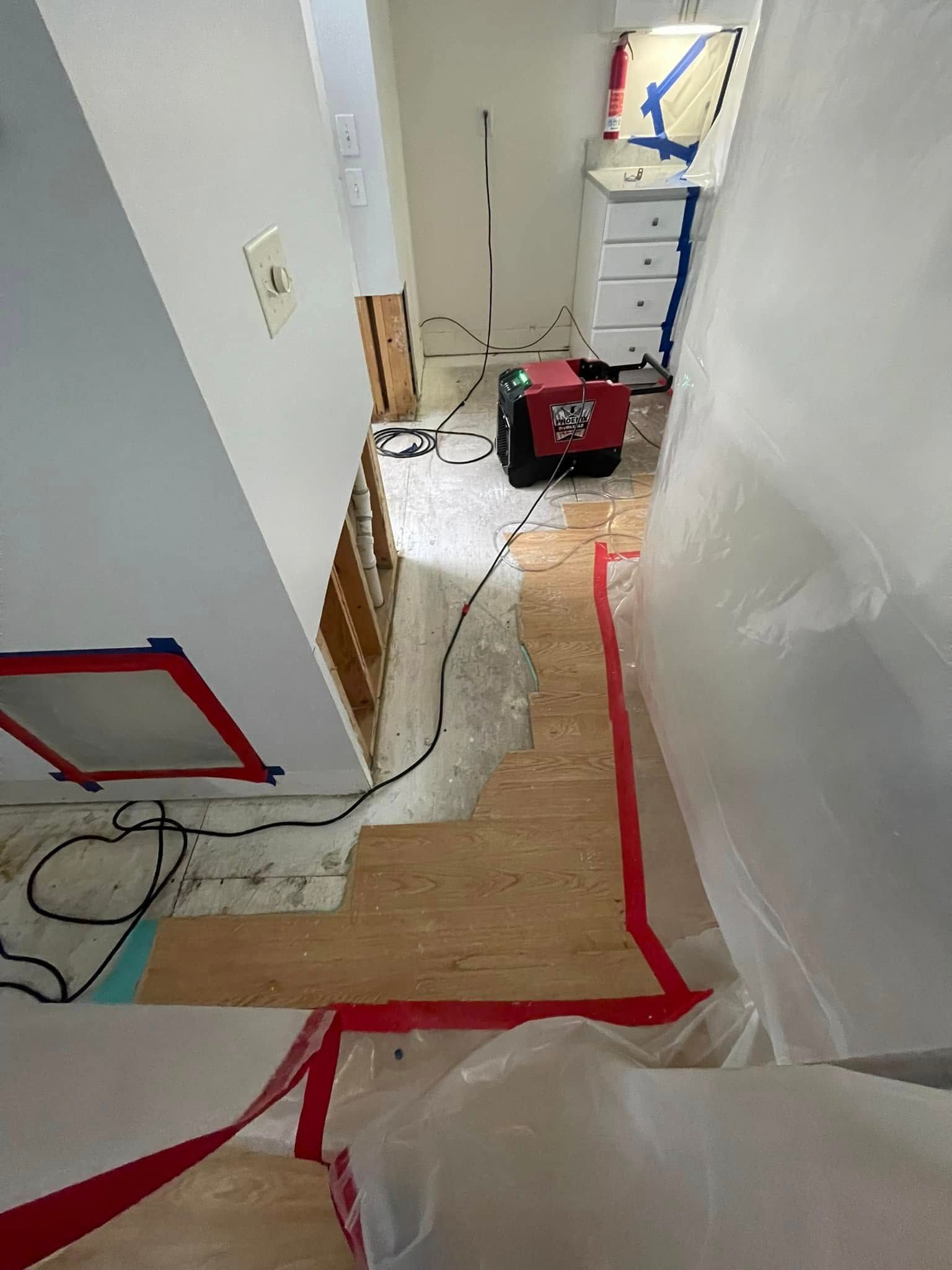 Interior view of a construction zone. Flooring removed; red tape on floor; equipment and plastic sheeting present.
