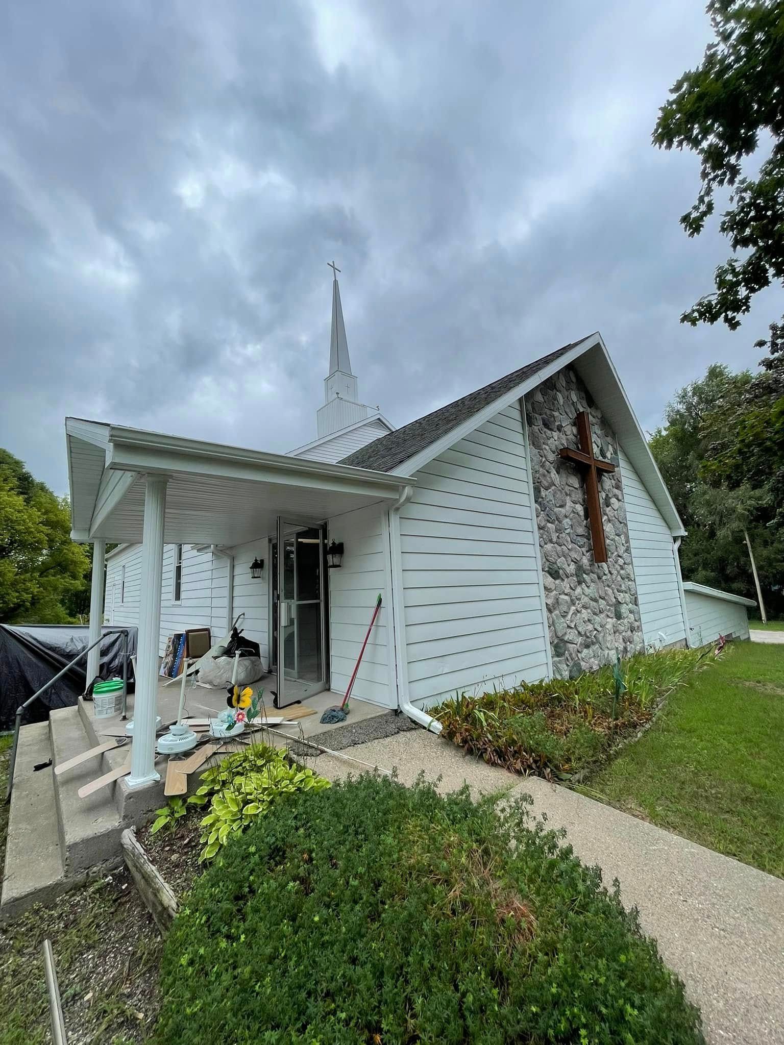 White church with steeple and stone cross, cloudy sky, green grass.
