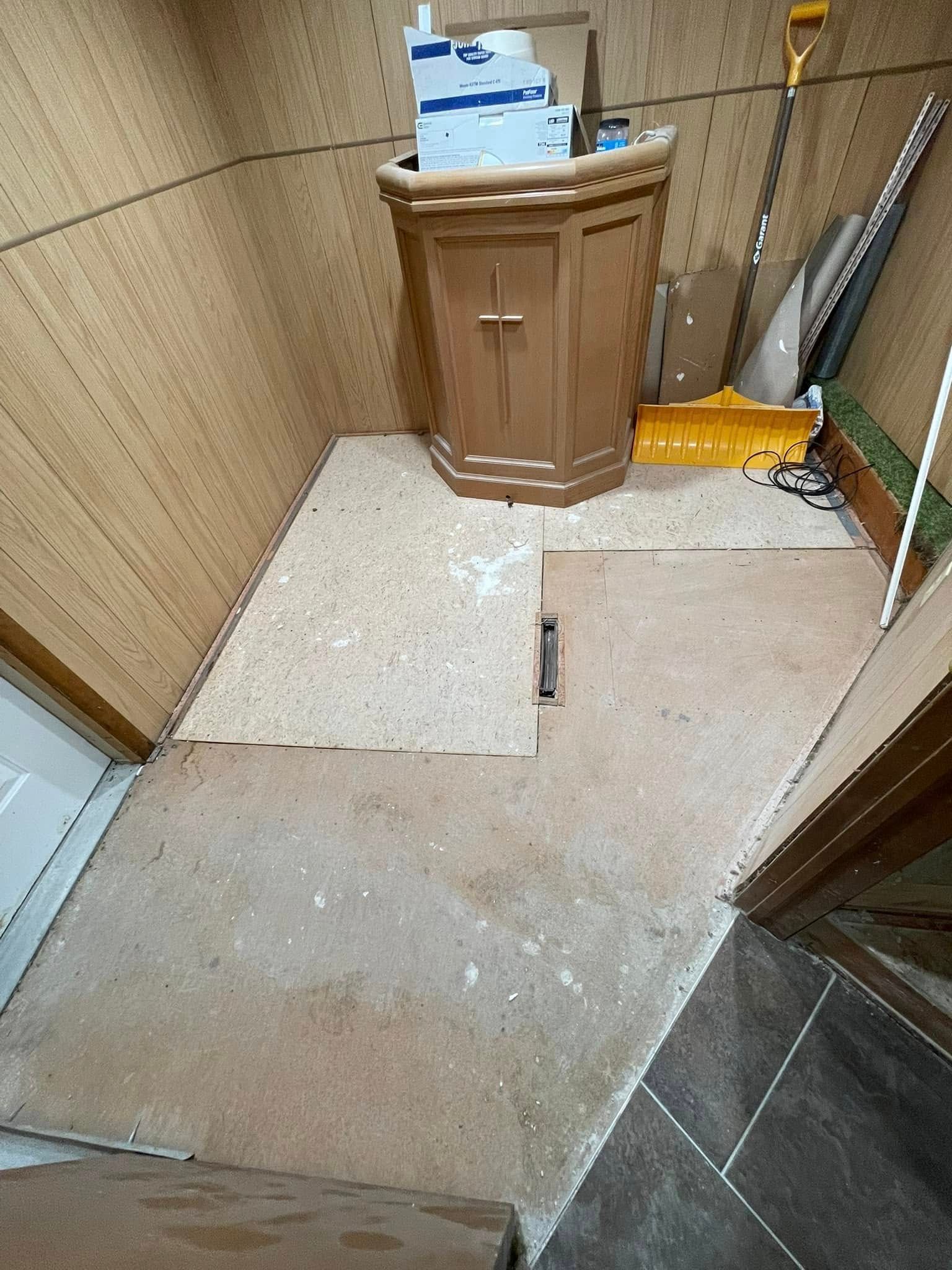 Wooden lectern in a room with unfinished floors and walls; tools and equipment stored in background.