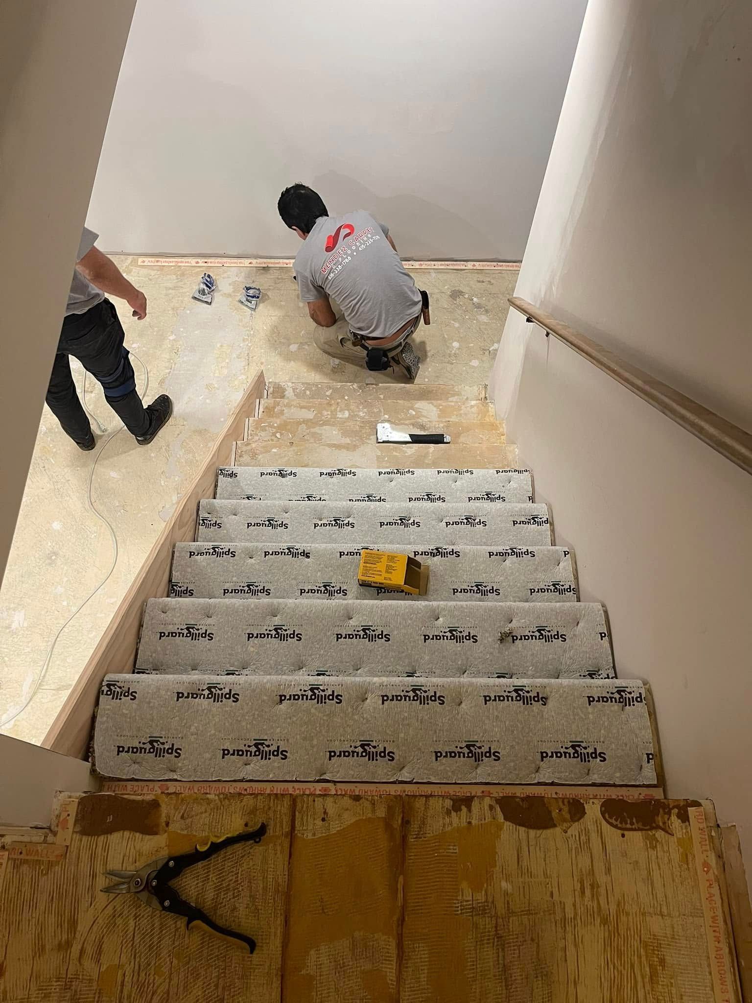 People installing carpet on a staircase, one kneeling. Staircase has partially installed carpet.