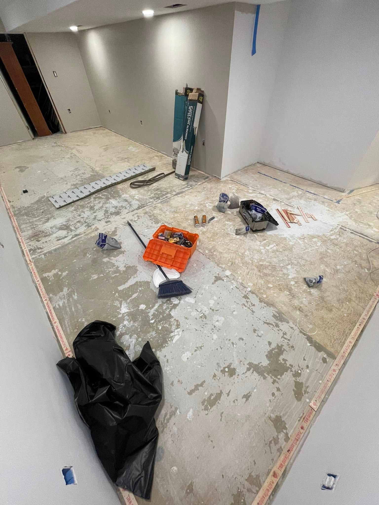Room under construction, with exposed floor, tools, and debris. Walls are white with a textured finish.