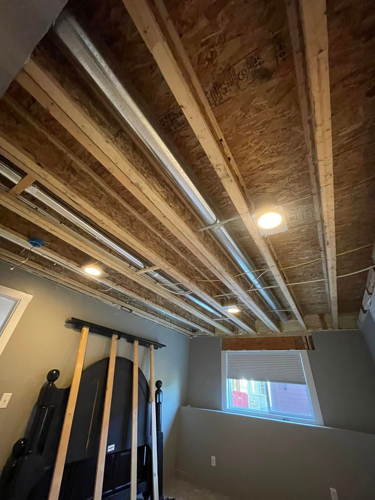 View of a room with exposed ceiling joists and recessed lighting; a bed and window are visible.