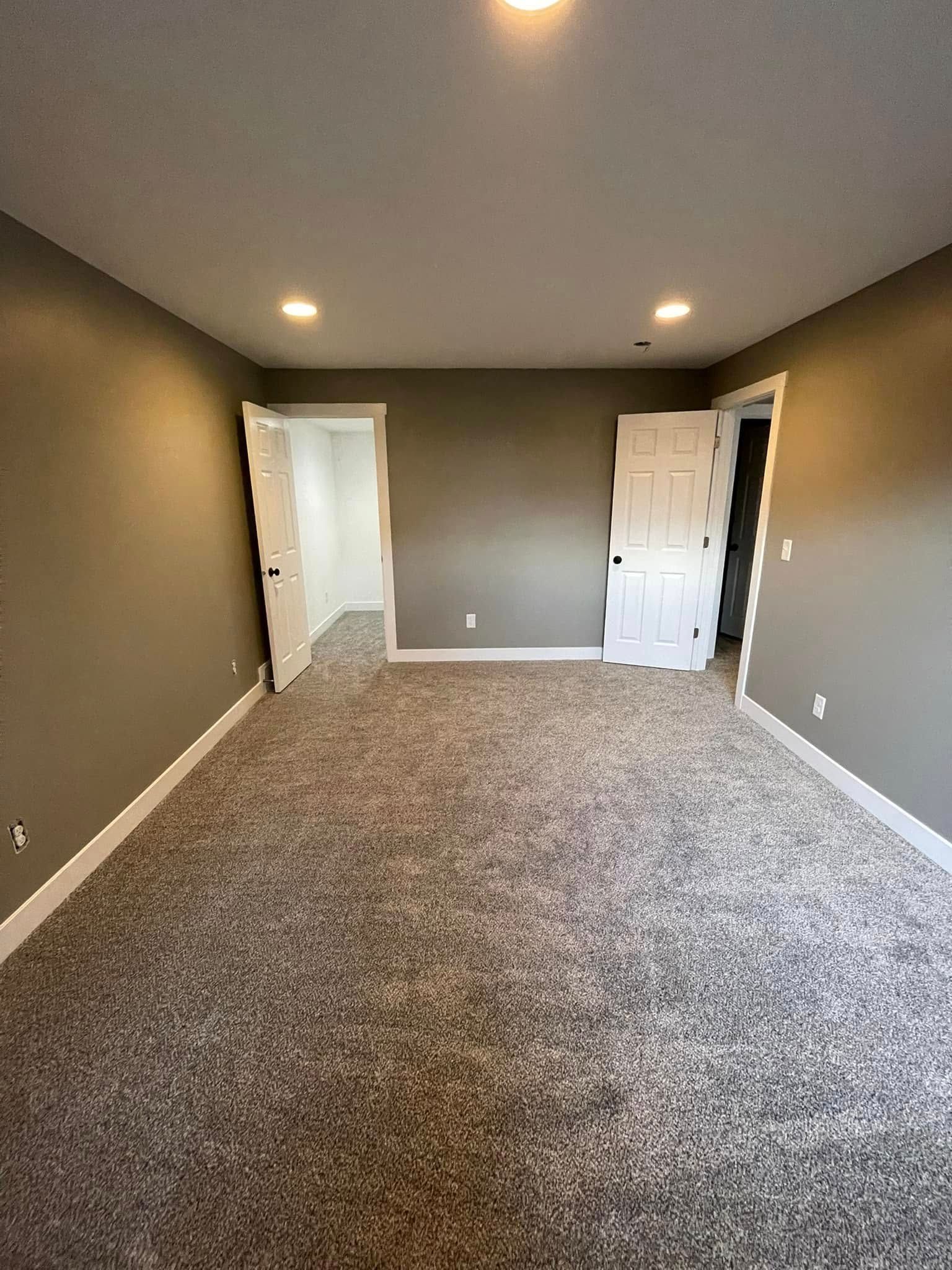 Empty room with gray walls, white trim, and speckled carpet; two doors and ceiling lights.